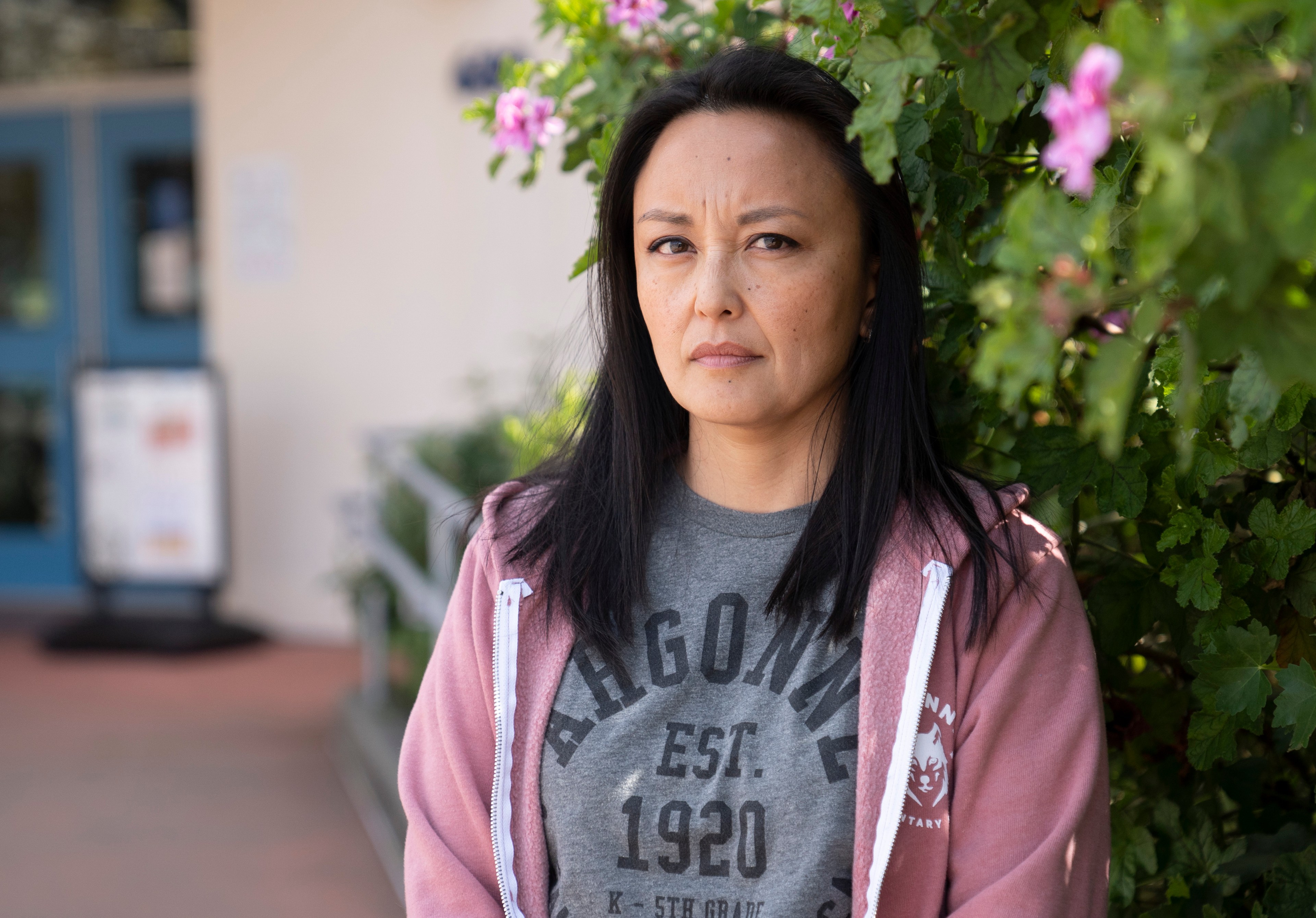 A woman stands outdoors, wearing a pink hoodie and gray t-shirt, looking serious. Behind her are green leaves and pink flowers, with a blue door in the background.