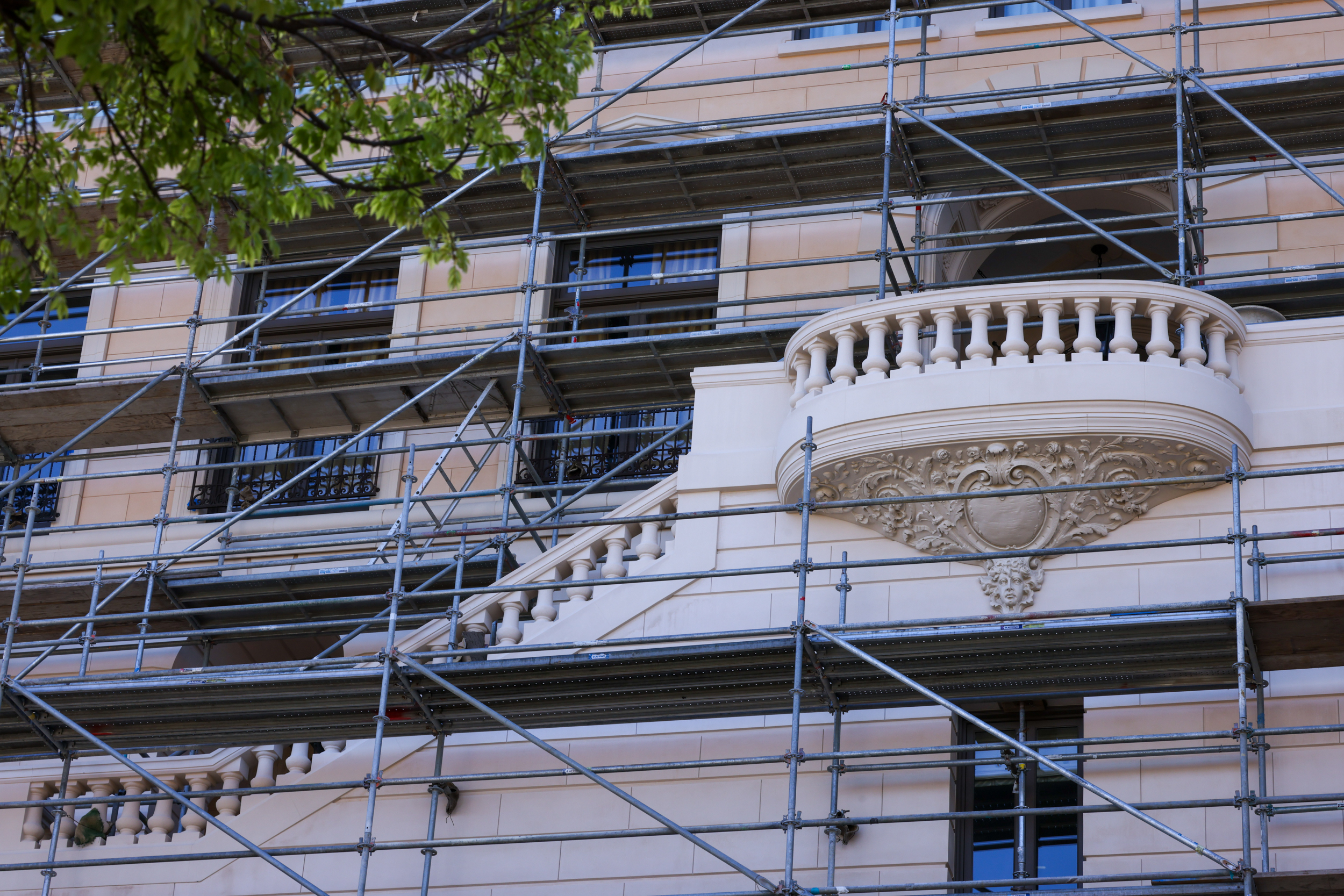 A building facade covered with scaffolding is shown, featuring intricate stonework and a small balcony. Leaves from a tree partially frame the top left.