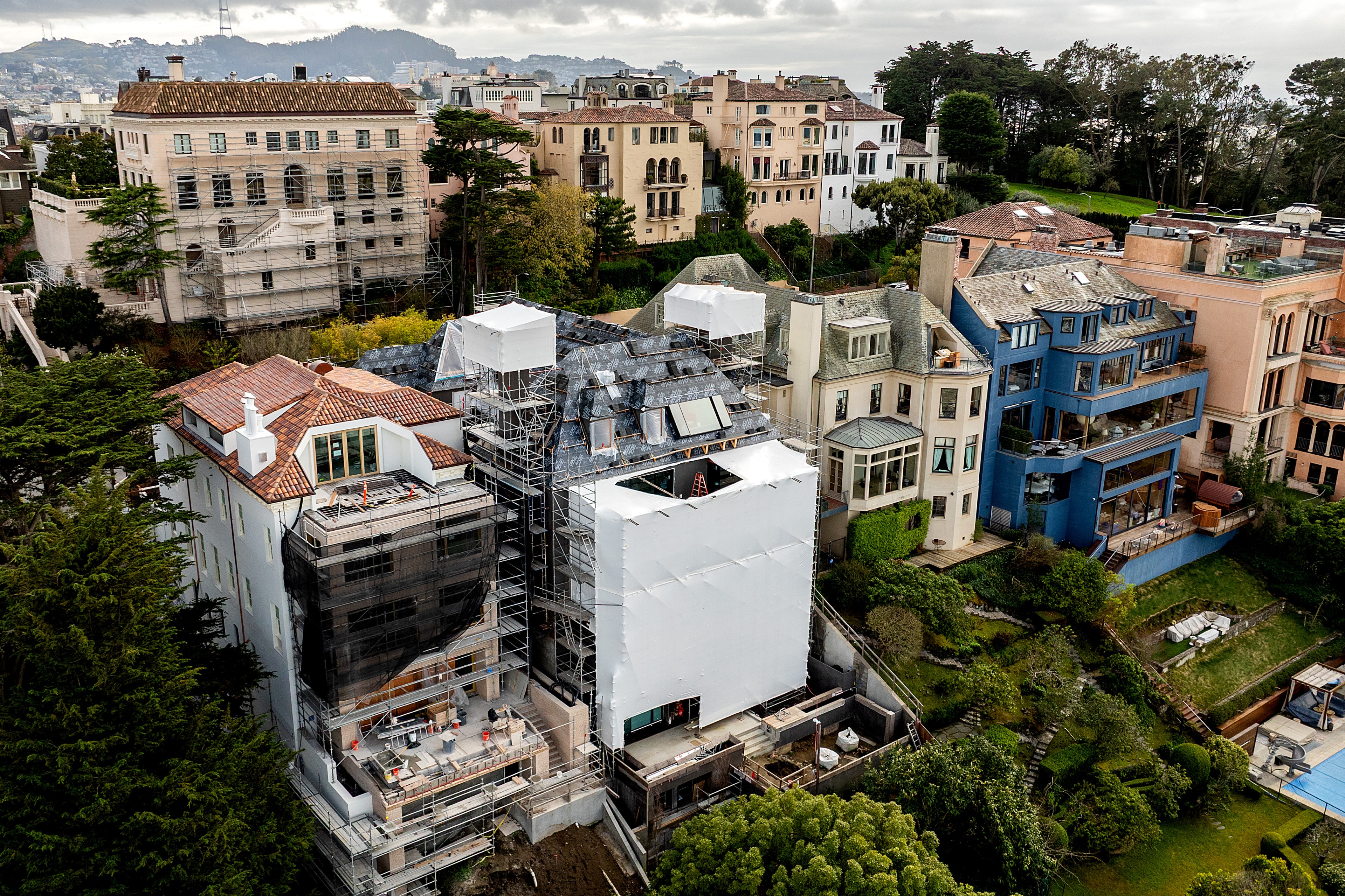 The image shows a cluster of large homes on a hillside, with one under renovation covered in scaffolding and white sheeting. Lush greenery surrounds the area.