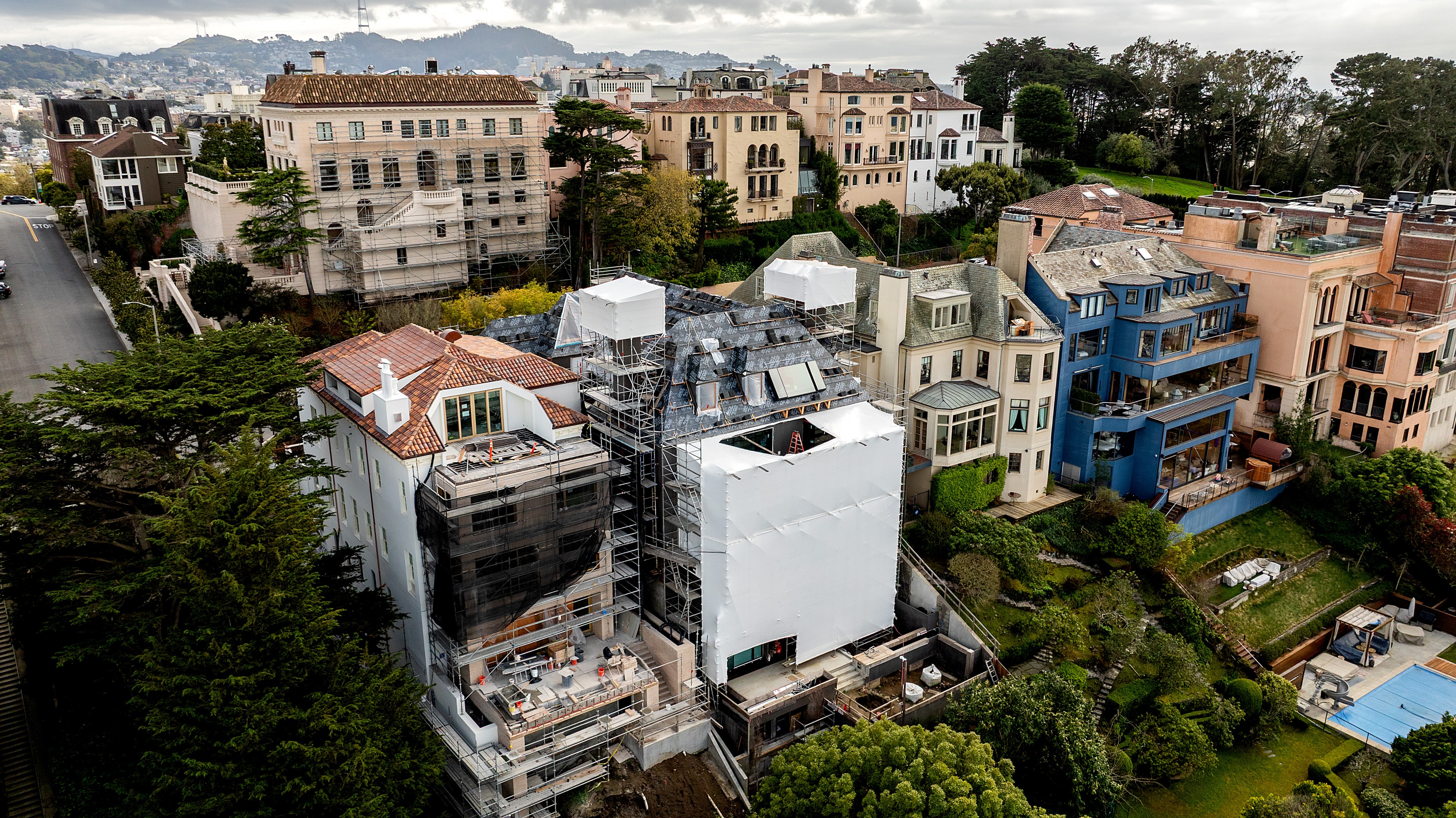 The image shows a cluster of large homes on a hillside, with one under renovation covered in scaffolding and white sheeting. Lush greenery surrounds the area.