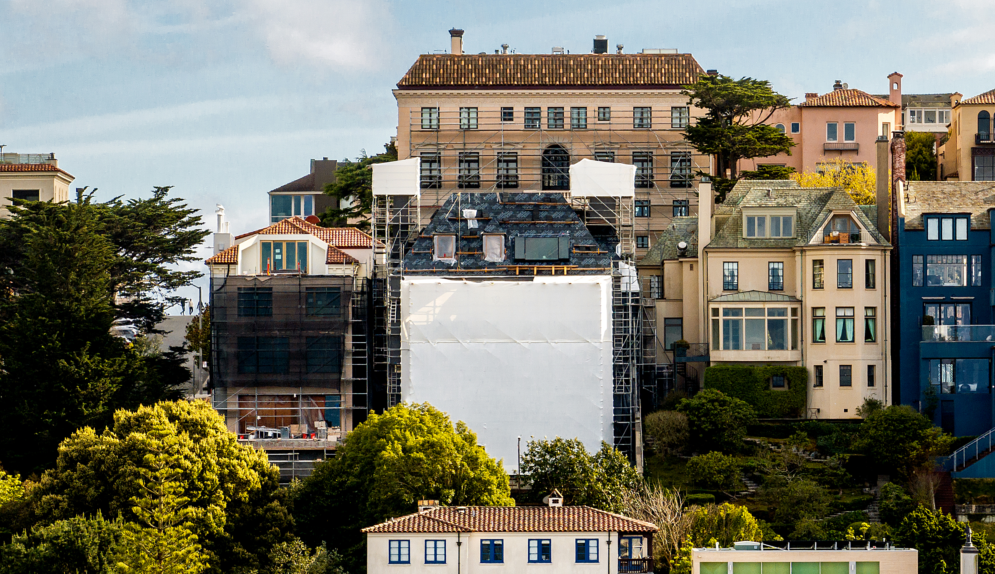 The image features a building covered with scaffolding and a large white tarp in front, surrounded by other colorful buildings and lush green trees.