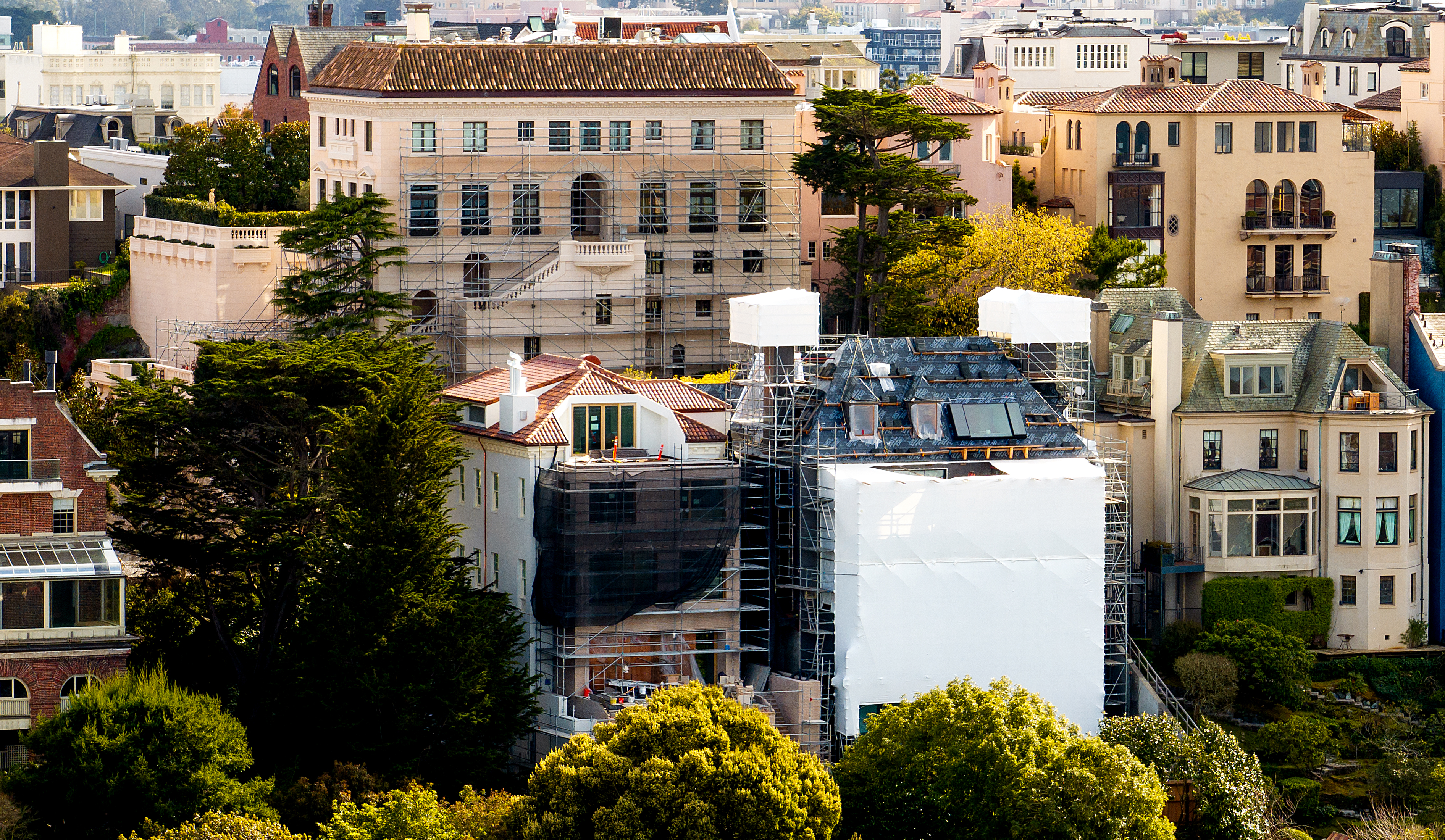 A densely packed neighborhood features historic multi-story buildings with scaffolding, surrounded by lush greenery and vibrant trees under a clear sky.