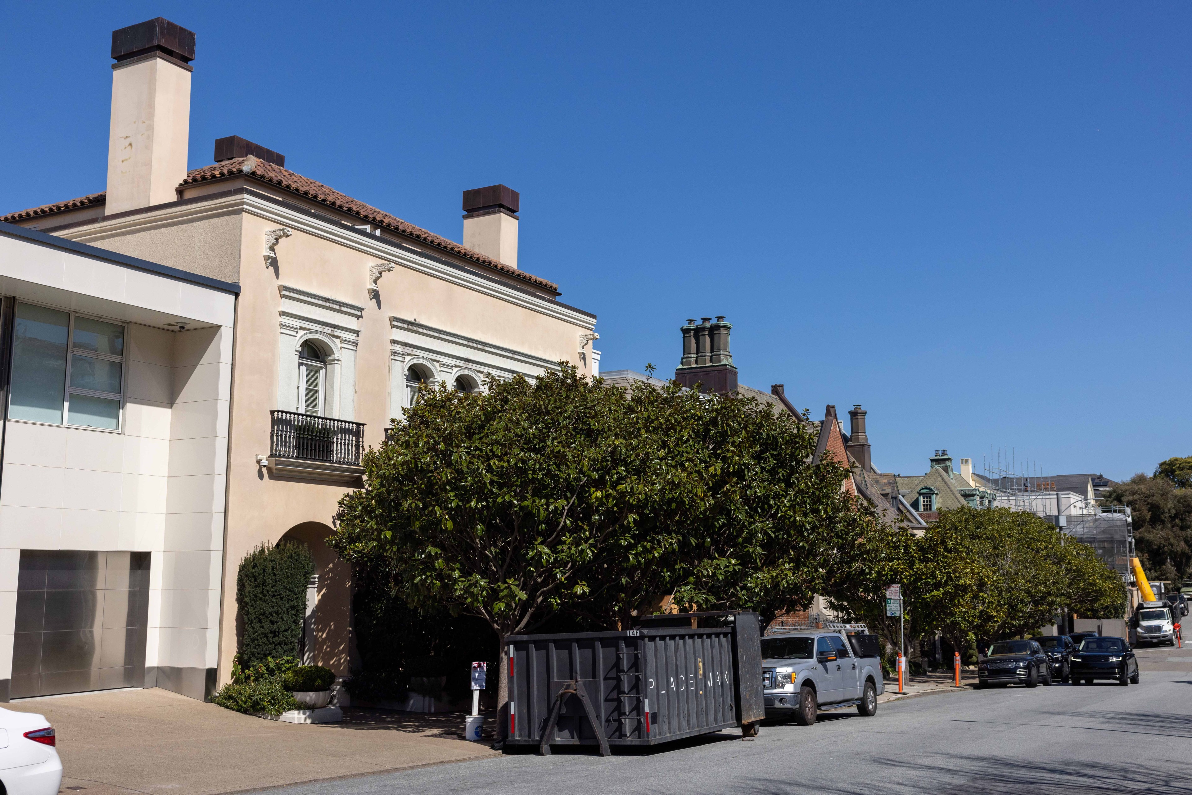 A street with a large beige building featuring chimneys and ornate windows. A tree partially hides a dumpster, and several cars are parked on the road.