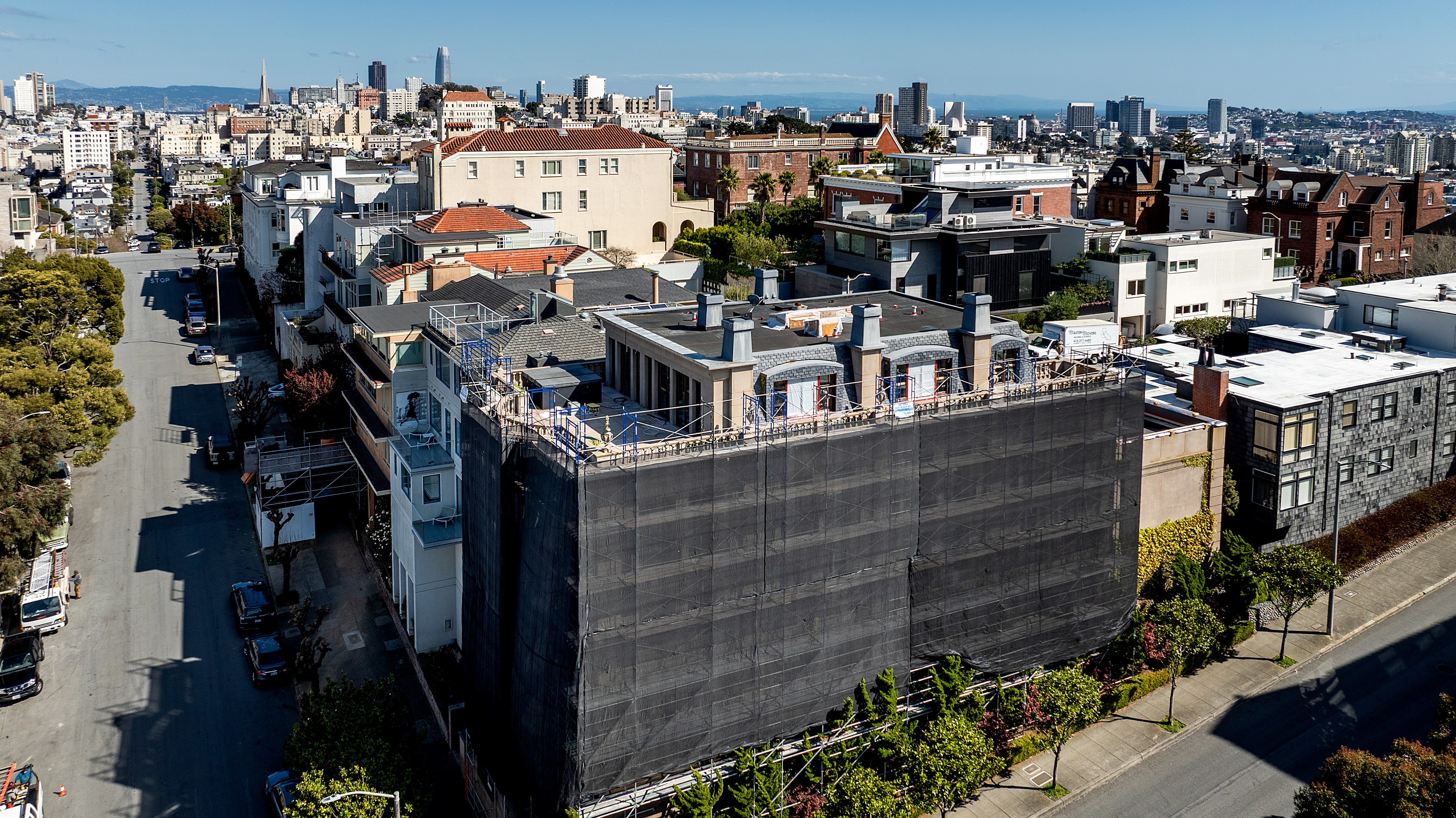 A cityscape shows a residential neighborhood with a prominent building covered in scaffolding and black netting, surrounded by various homes under a clear sky.