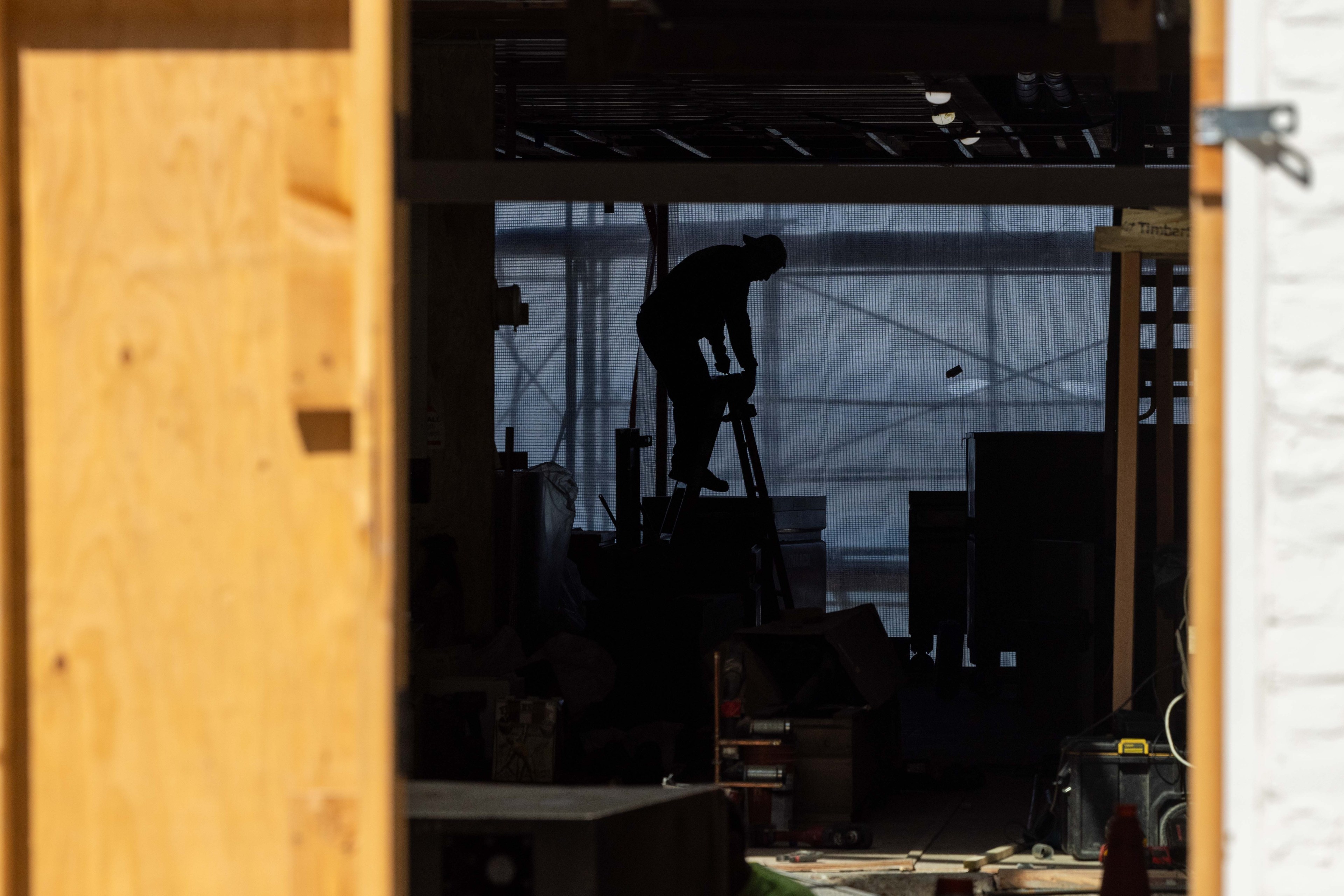 A silhouette of a person stands on a ladder inside a dimly lit construction site, framed by wooden beams and scattered tools.