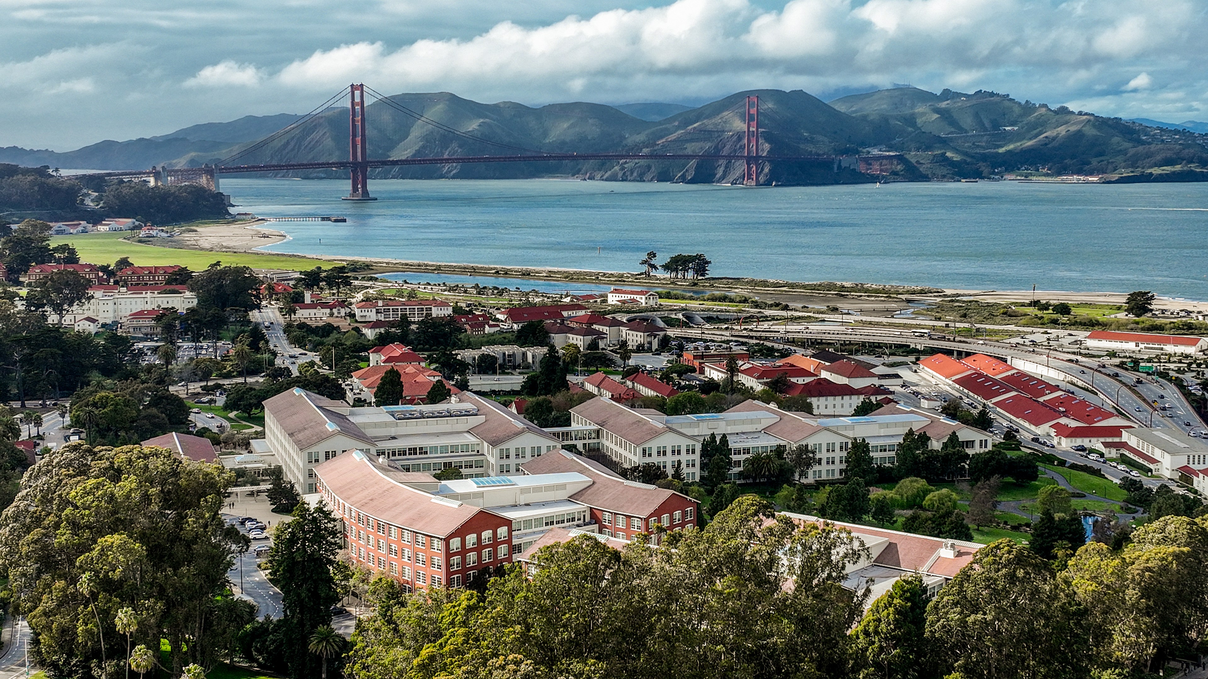 The image shows a scenic view of the Golden Gate Bridge over a large body of water. In the foreground, there are buildings with red roofs surrounded by lush greenery.