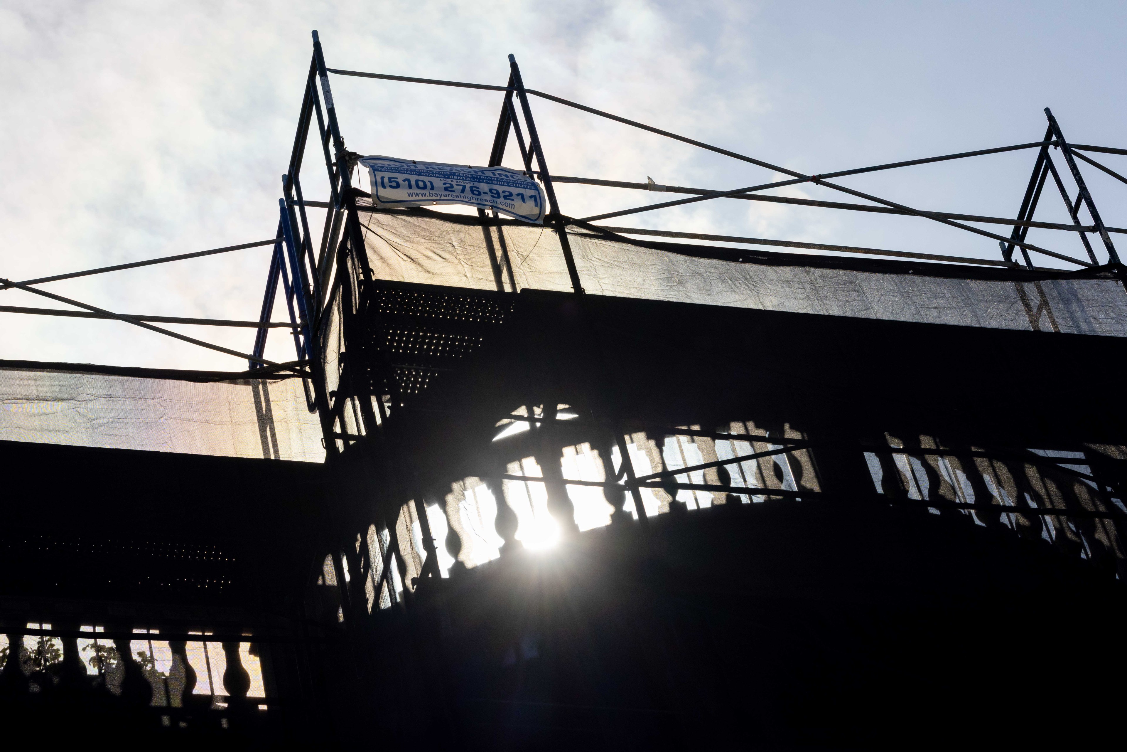 The image shows silhouetted scaffolding against a bright sky with the sun peeking through. A sign with a phone number is visible at the top.