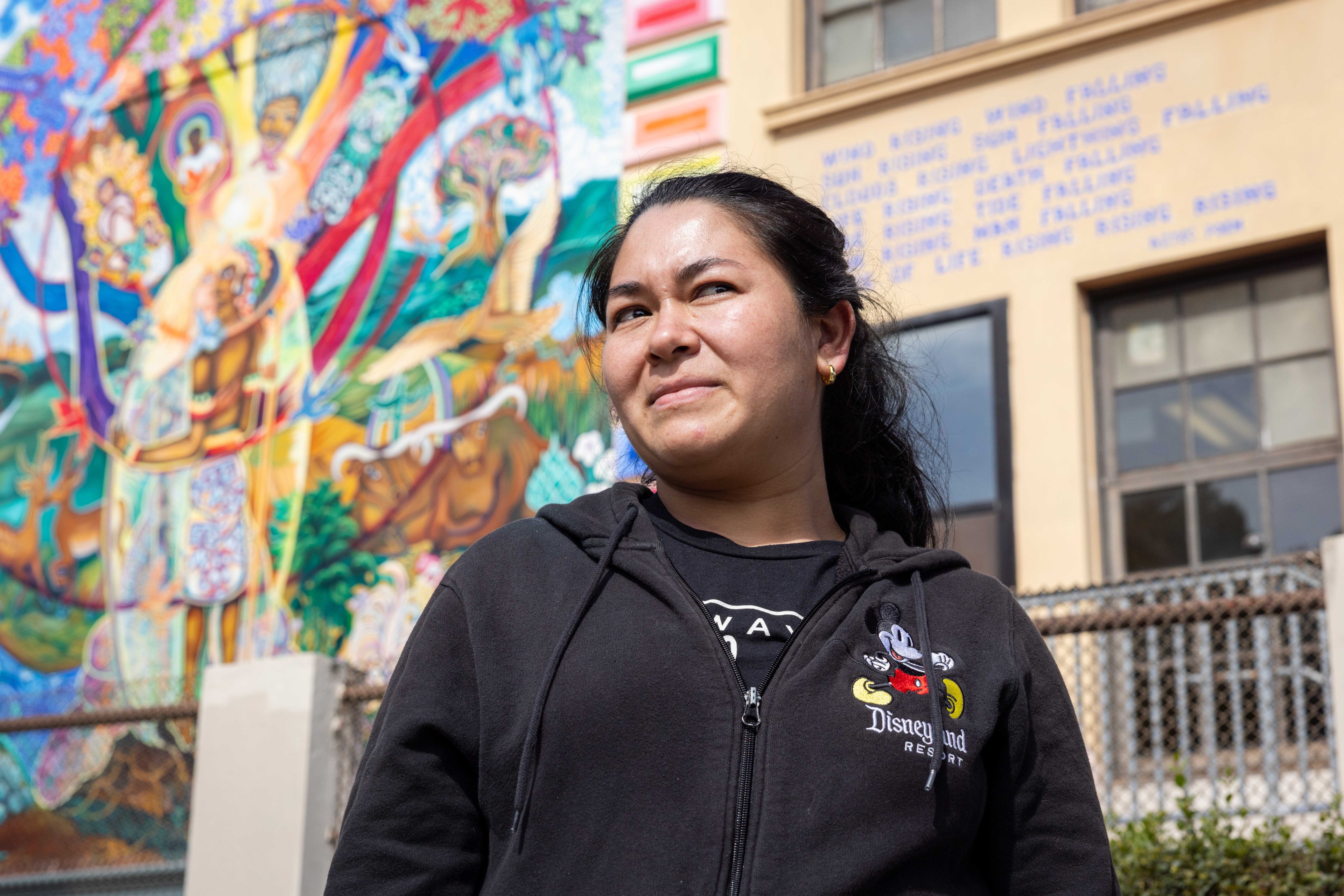 A person wearing a black Disneyland hoodie stands in front of a colorful mural depicting various figures and animals. A poem is visible on a building wall.