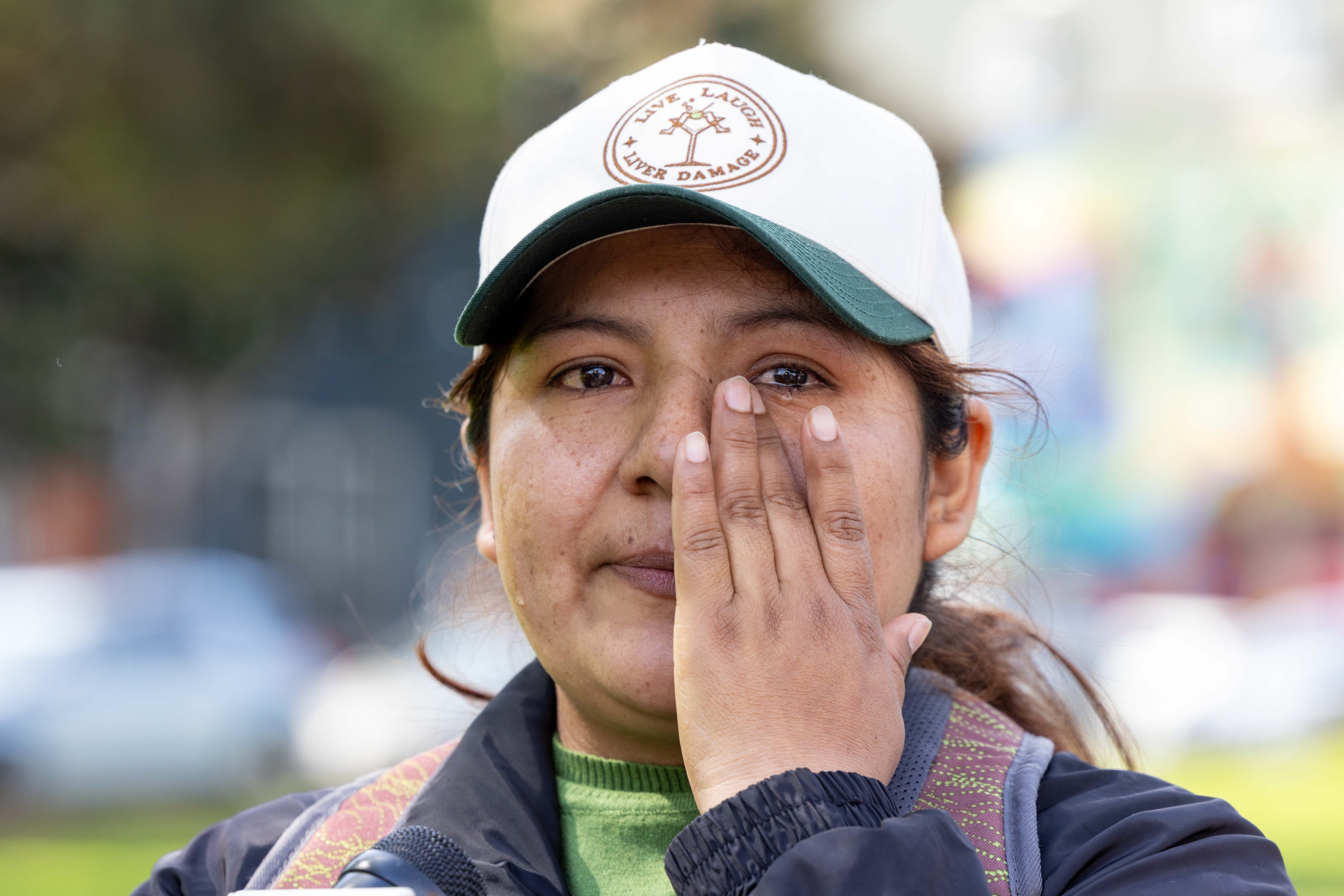 A person in a white and green cap is outdoors. The cap has a logo with the text "Live, Laugh, Liver Damage." The person is touching their face with one hand.