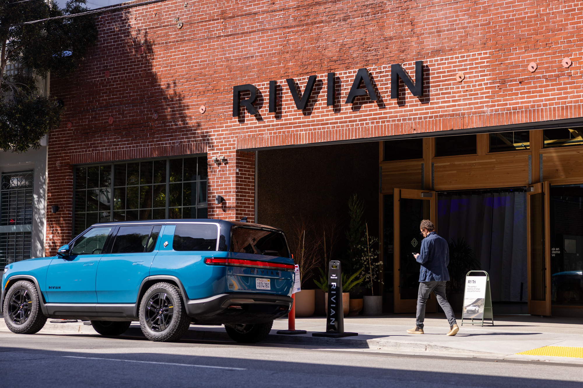 A blue Rivian SUV is parked in front of a brick building with a Rivian sign. A person walks nearby, looking at their phone.