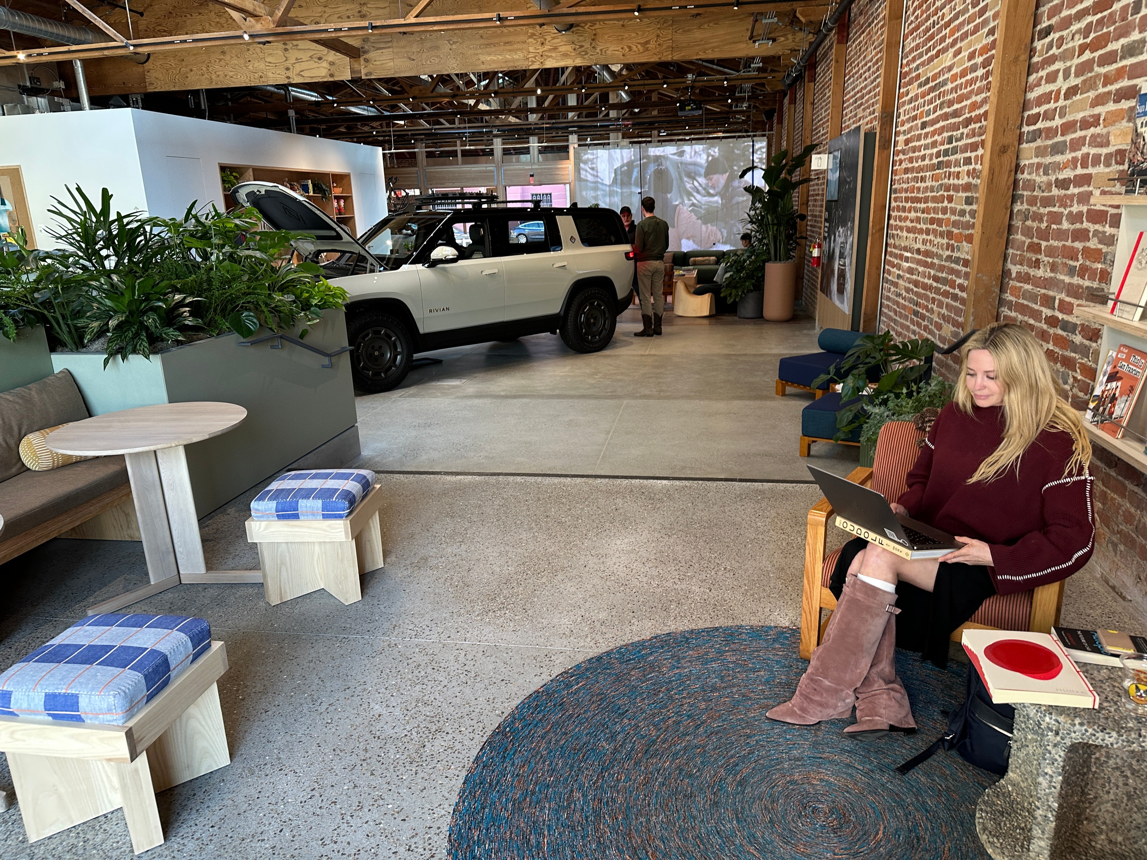 A woman sits on a chair using a laptop in a modern, plant-filled room with exposed brick. A white SUV with its trunk open is parked inside, with people nearby.