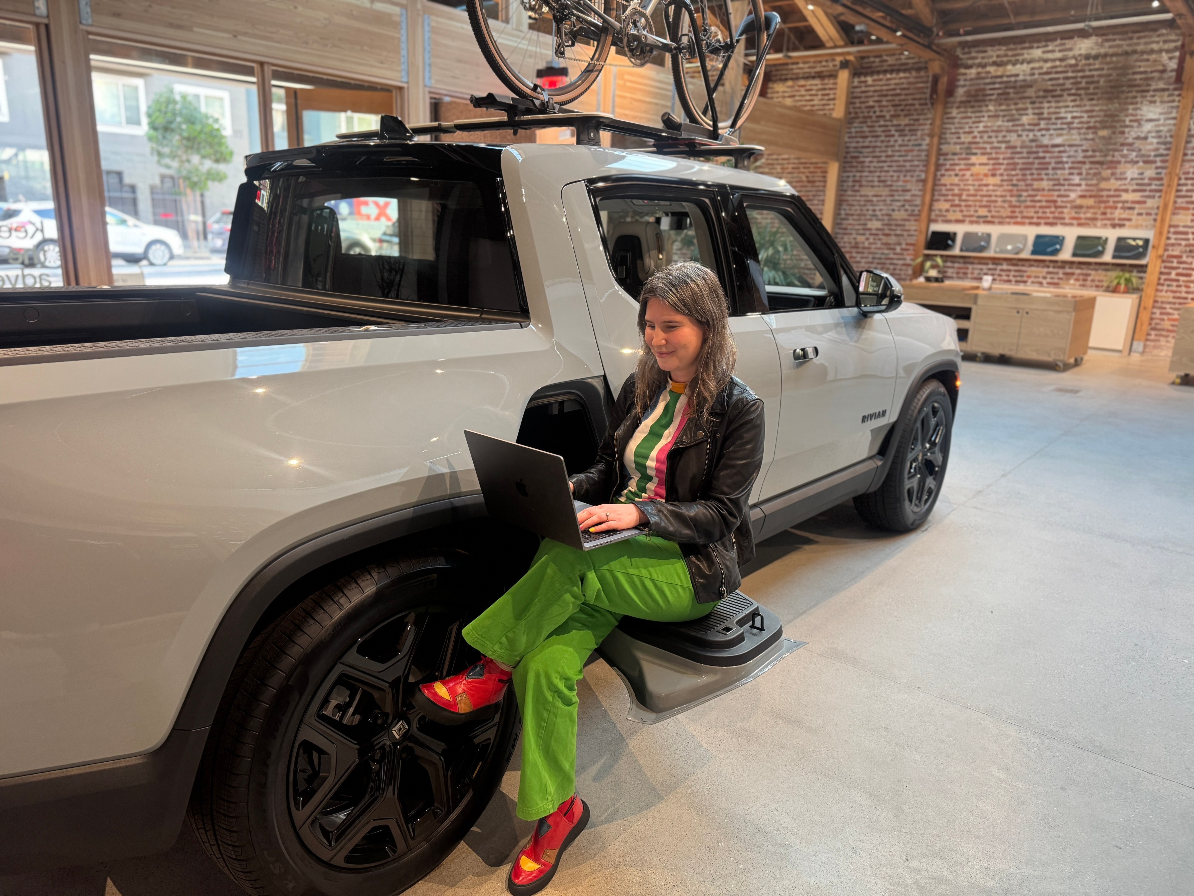 A woman in colorful attire sits on a truck's sidestep, using a laptop. The truck is indoors with bicycles on its roof and a brick wall in the background.