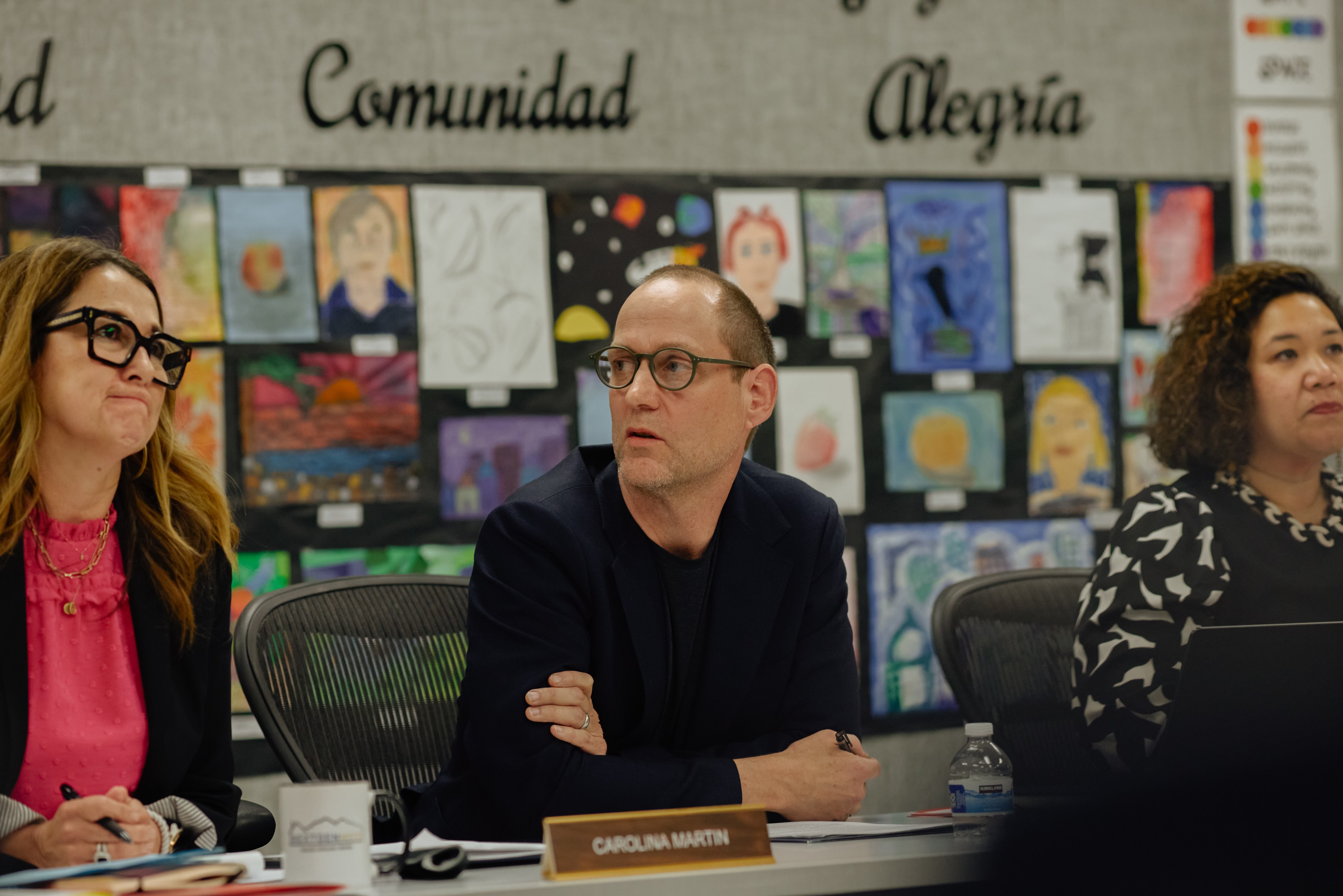 Three people sit at a table with colorful artwork on the wall behind them. They are focused on something ahead, with a nameplate reading &quot;Carolina Martin.&quot;