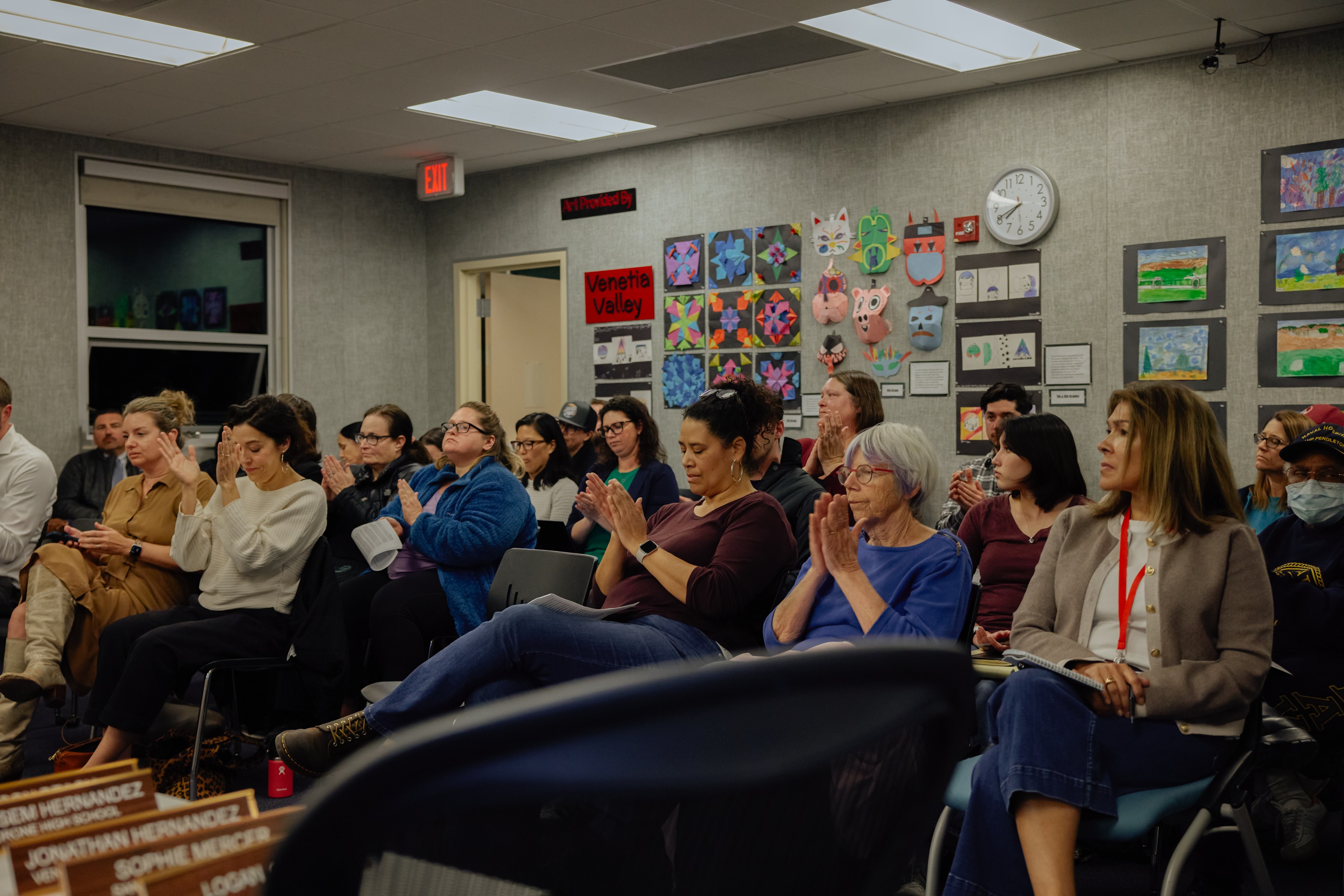 A group of people is seated in a room, attentively clapping. Artworks are displayed on the walls, and a clock shows the time as 10:12.