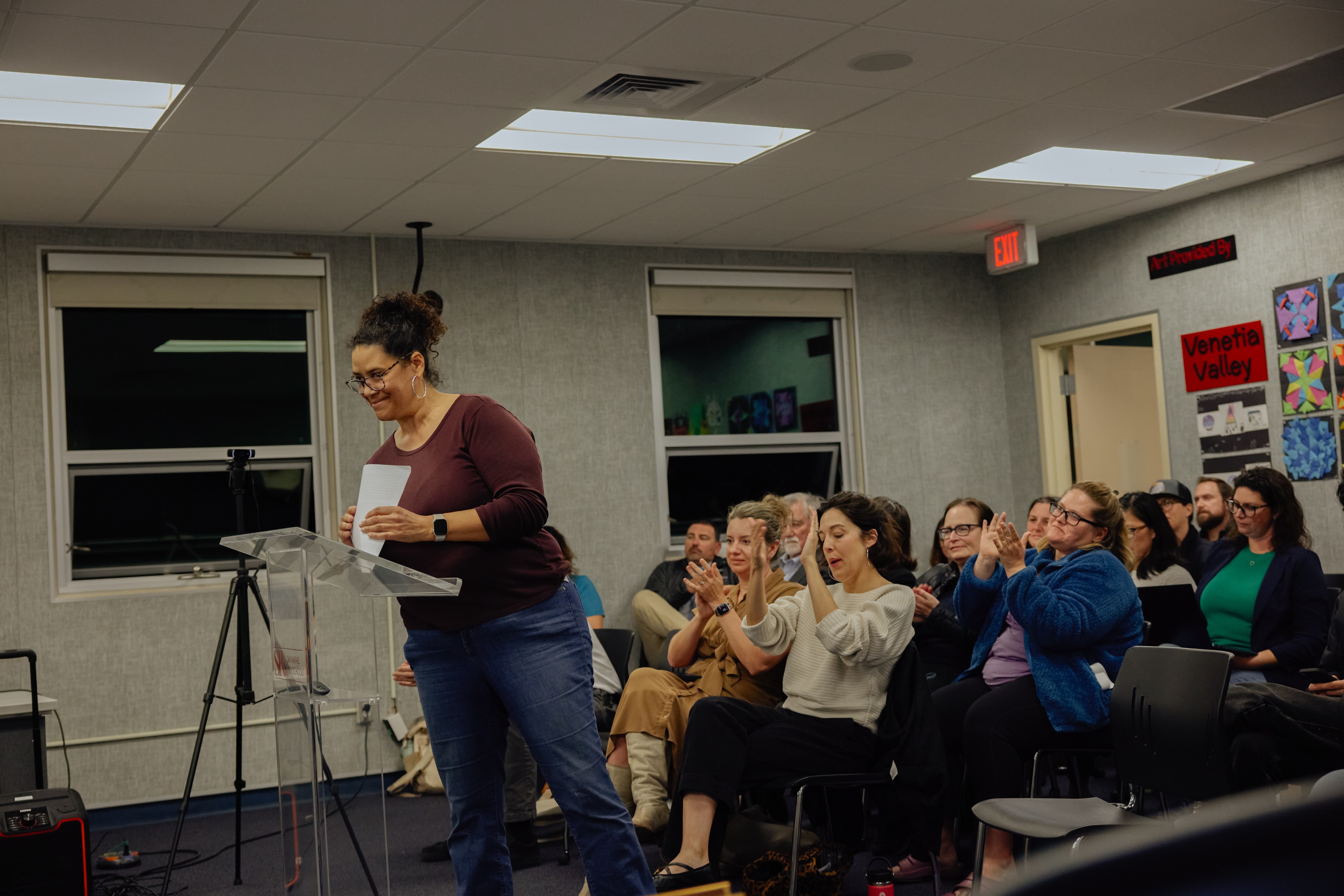 A woman stands at a podium holding papers, smiling, while an audience in a room with artworks and a sign claps and watches her attentively.