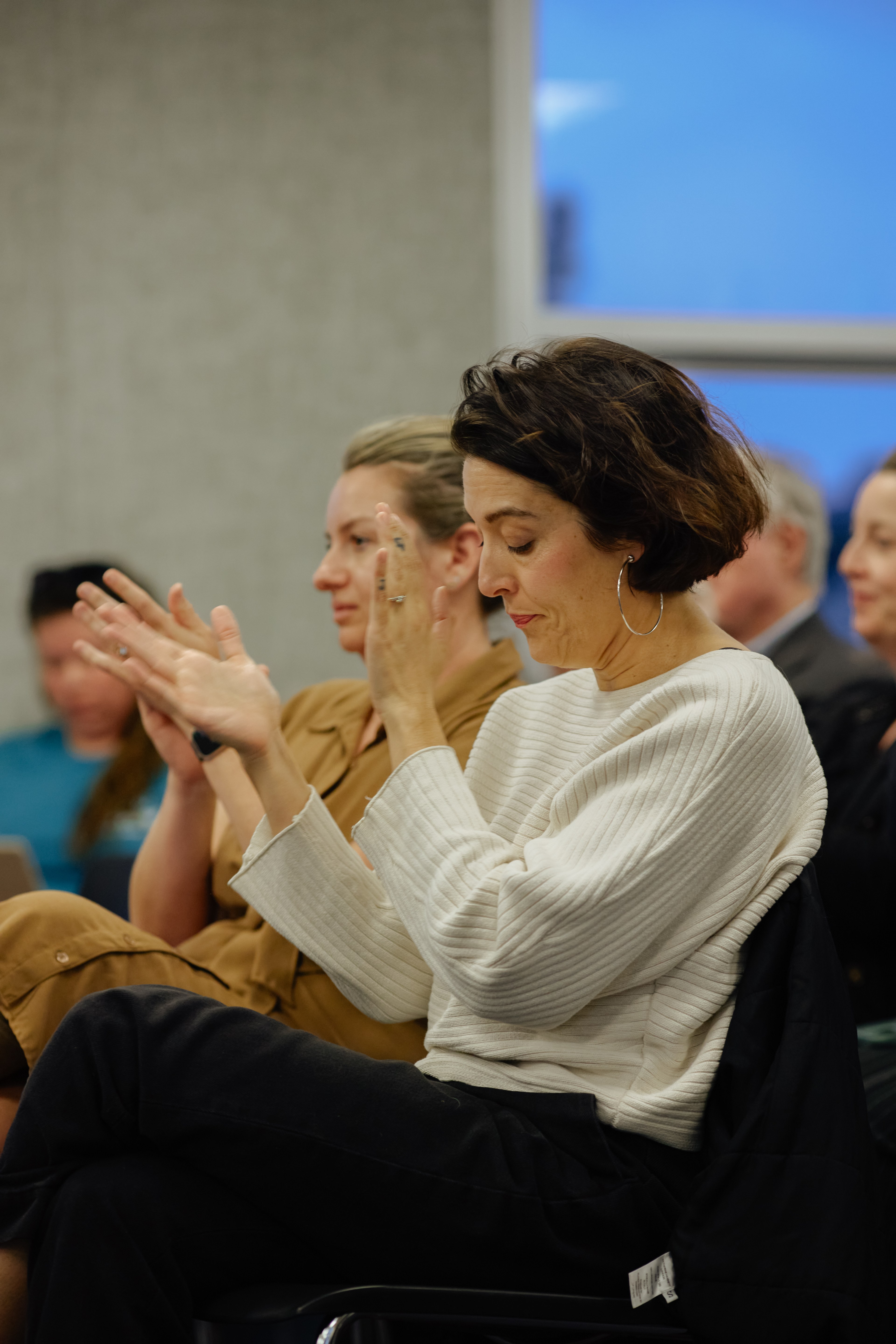 A group of people, mainly women, are sitting and applauding. The focus is on a woman wearing a white sweater with short dark hair, clapping her hands.