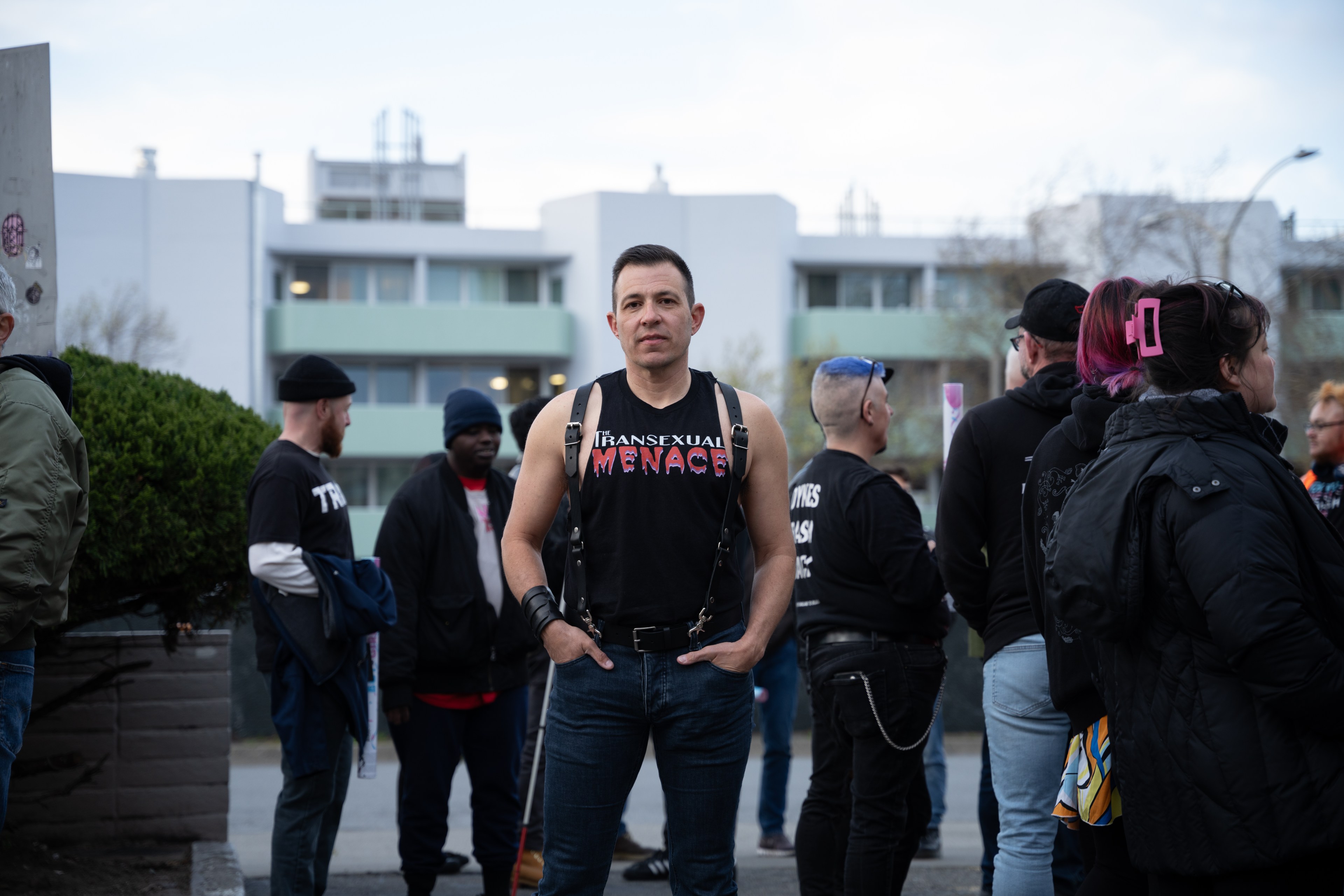 A person stands confidently in a sleeveless shirt that reads "Transsexual Menace," surrounded by a casually gathered group, in an outdoor urban setting.