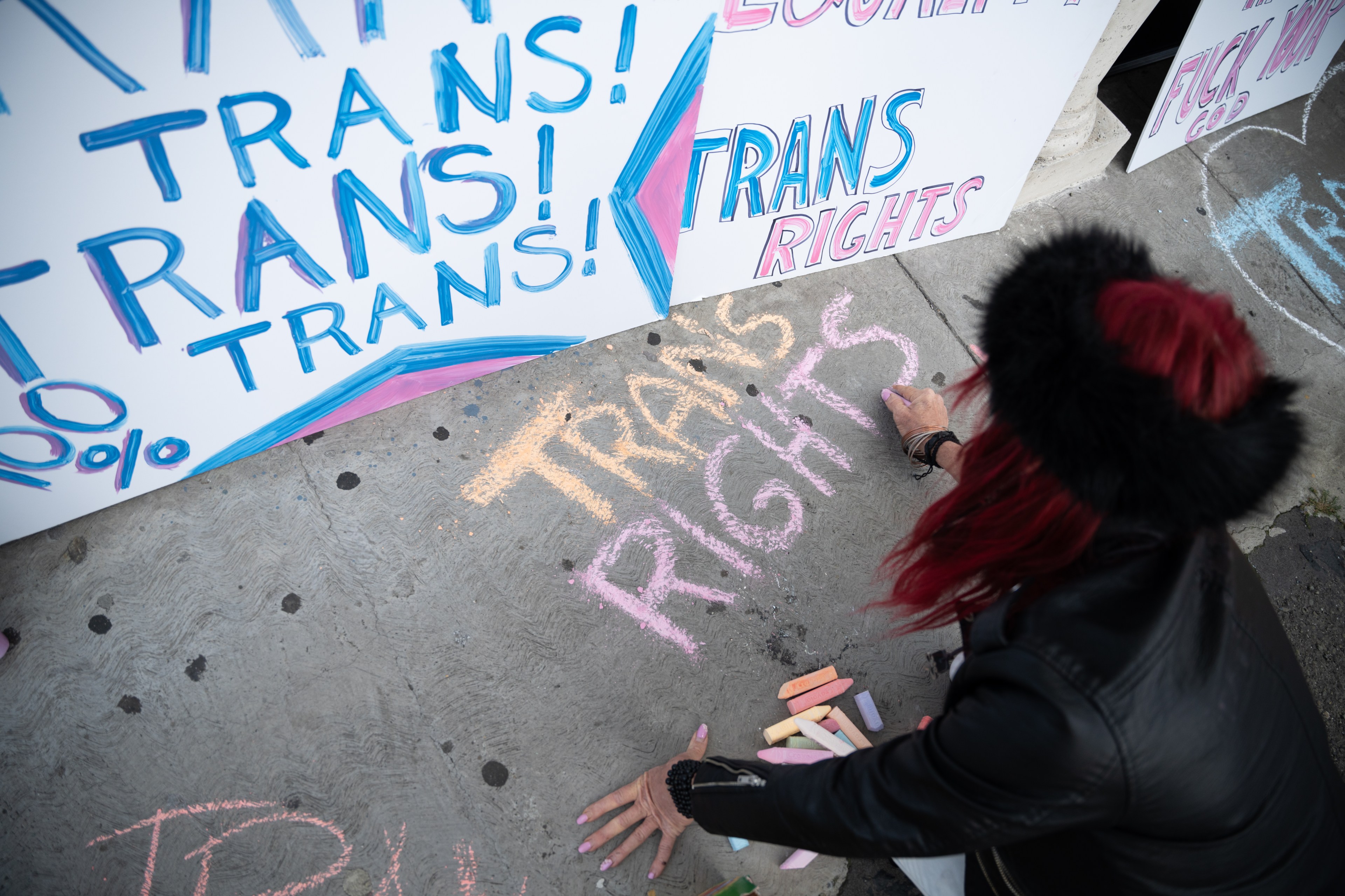 A person with red hair writes &quot;TRANS RIGHTS&quot; in colorful chalk on the ground near posters with &quot;TRANS!&quot; and &quot;TRANS RIGHTS&quot; text in blue and pink.