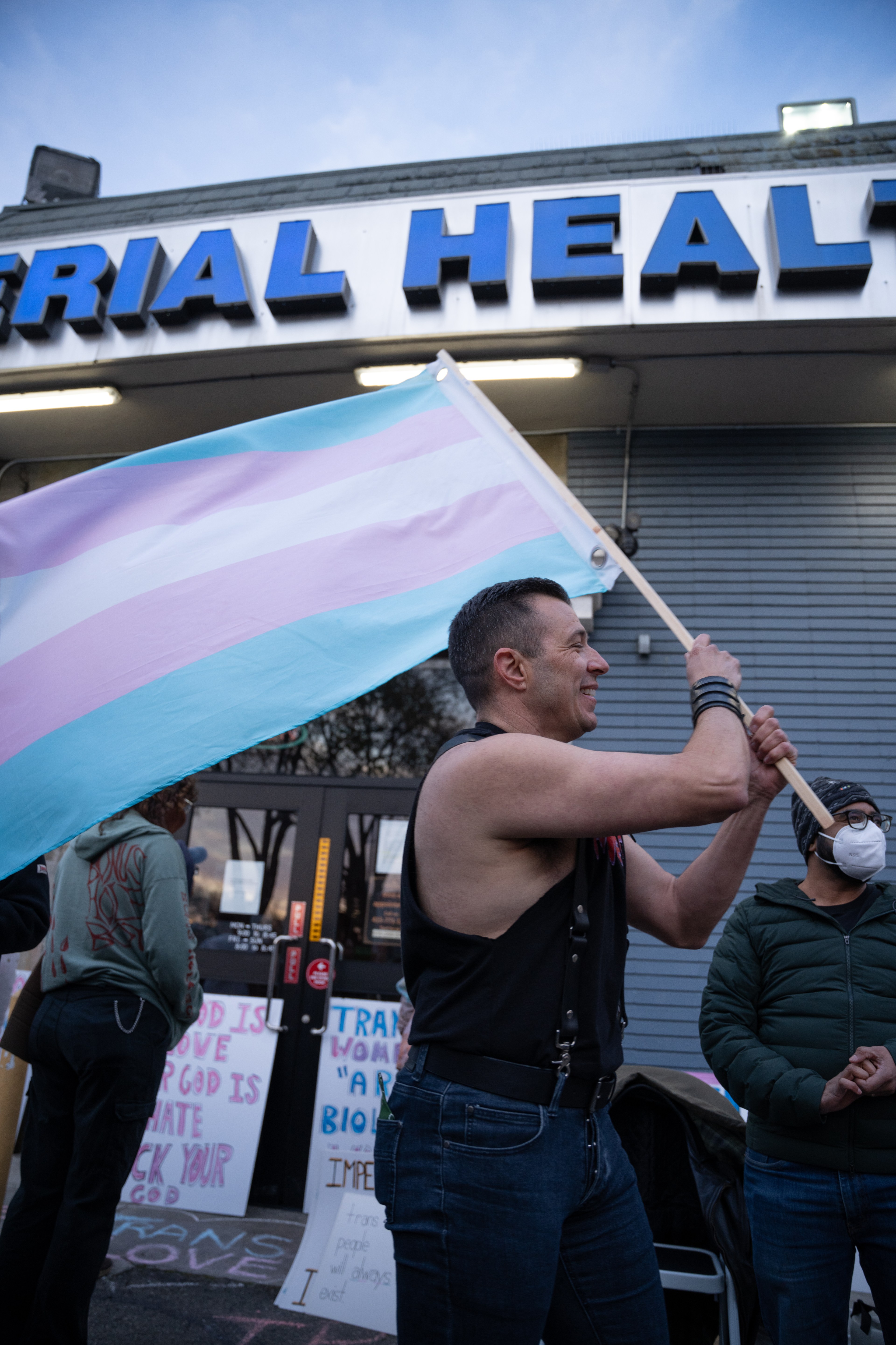 A person proudly holds a transgender pride flag outside a building with banners advocating for transgender rights.