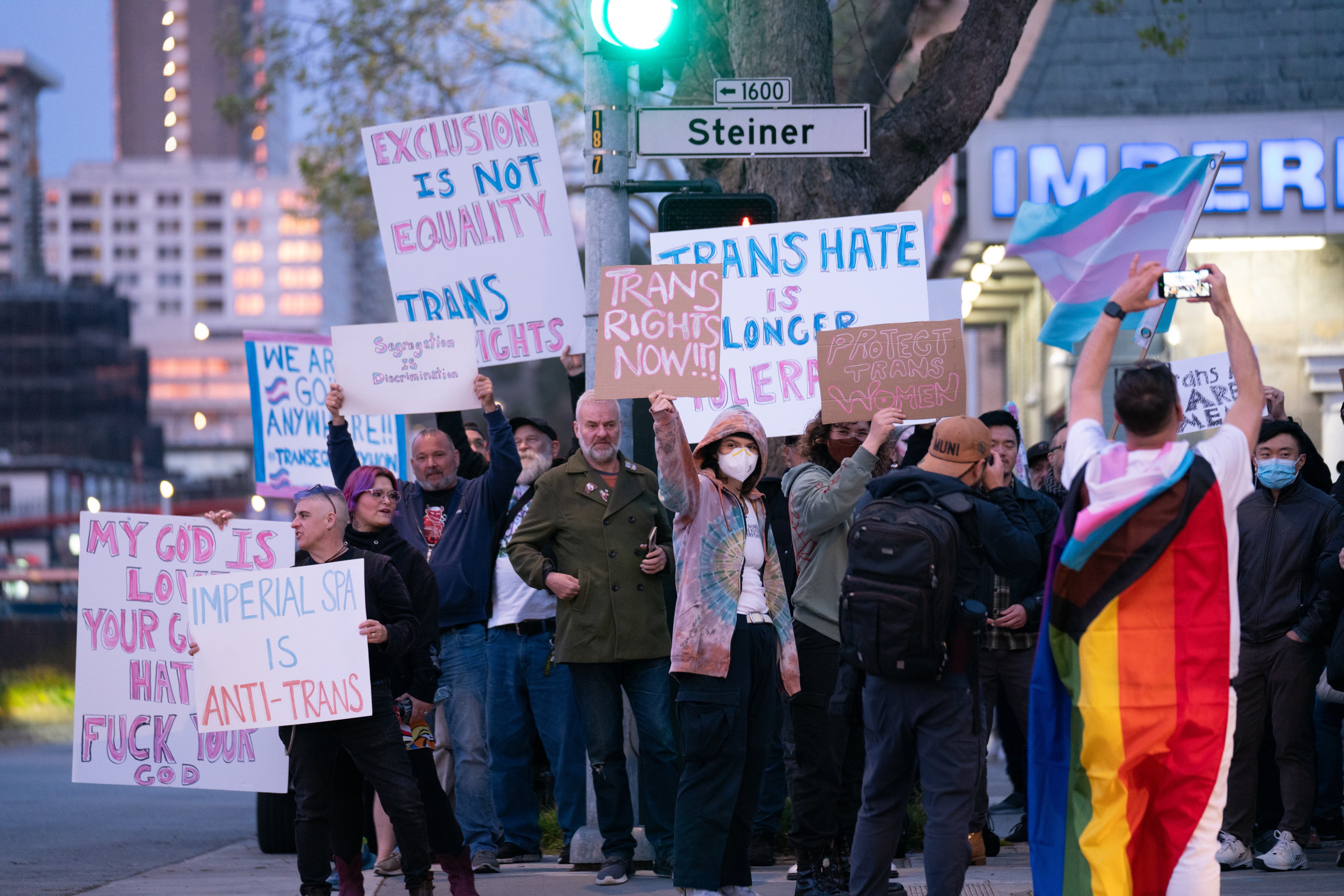 A group of people holds signs advocating for trans rights. One wears a pride flag, and a trans flag is visible. They stand under a street sign in an urban area.