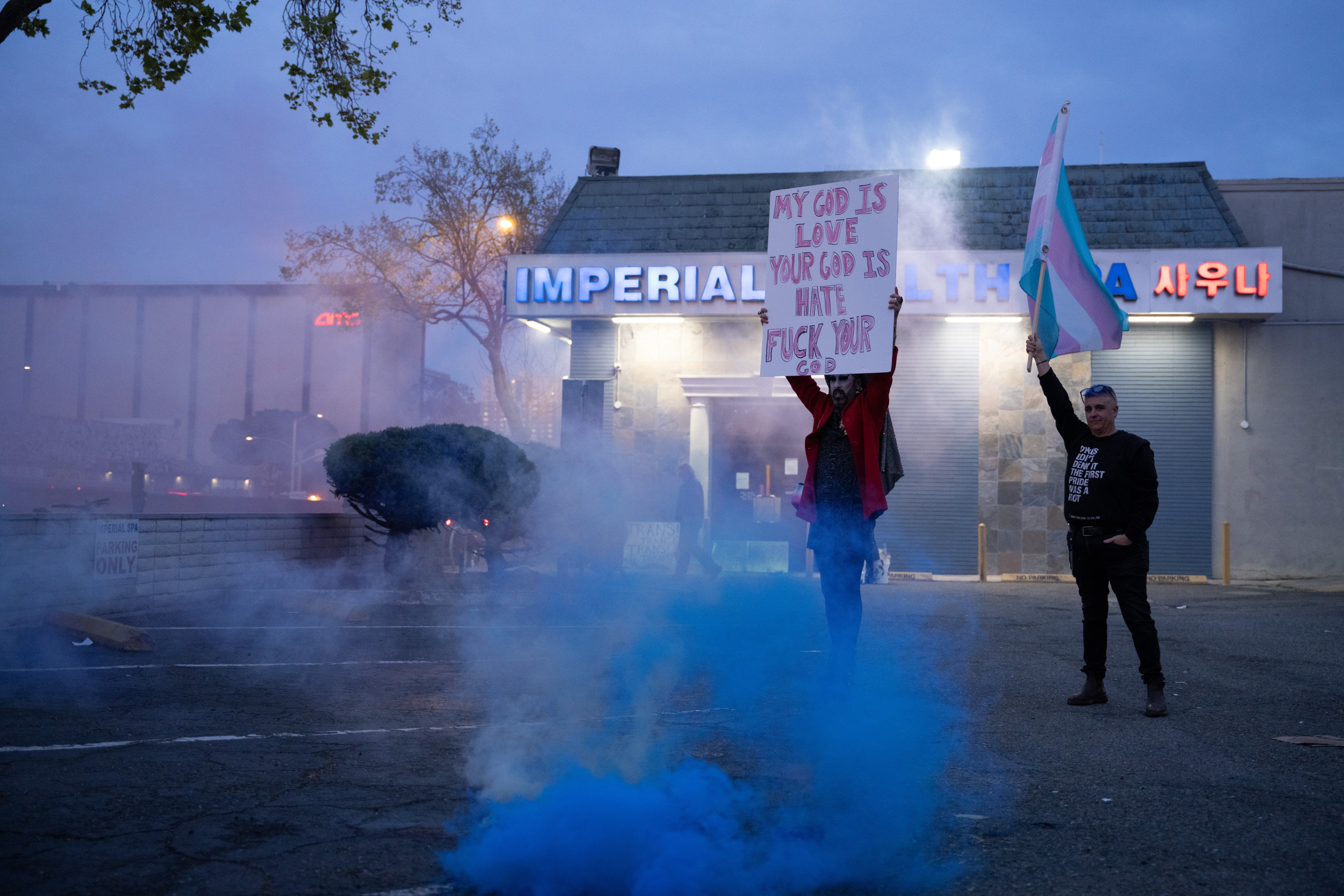 Two people stand amid blue smoke outside a building, holding a sign and a trans flag. A neon sign reads &quot;IMPERIAL HEALTH SPA.&quot;