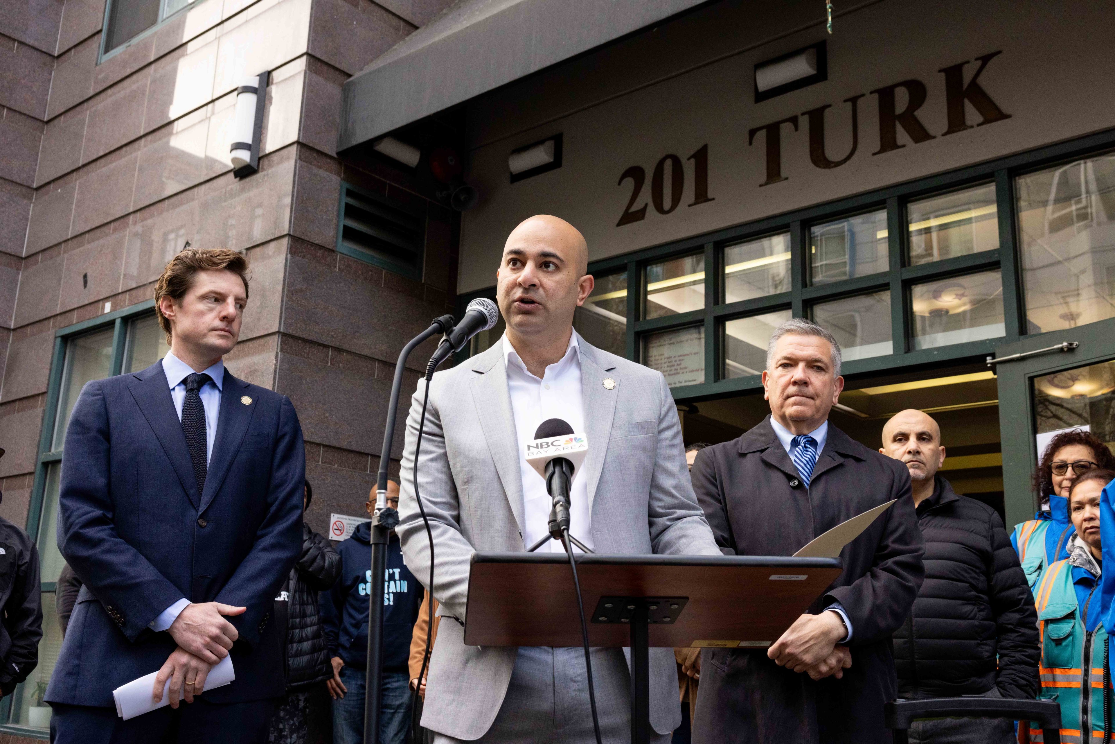 A man is speaking at a podium with a microphone labeled &quot;NBC&quot; outside a building marked &quot;201 Turk.&quot; Several people are standing nearby, listening attentively.