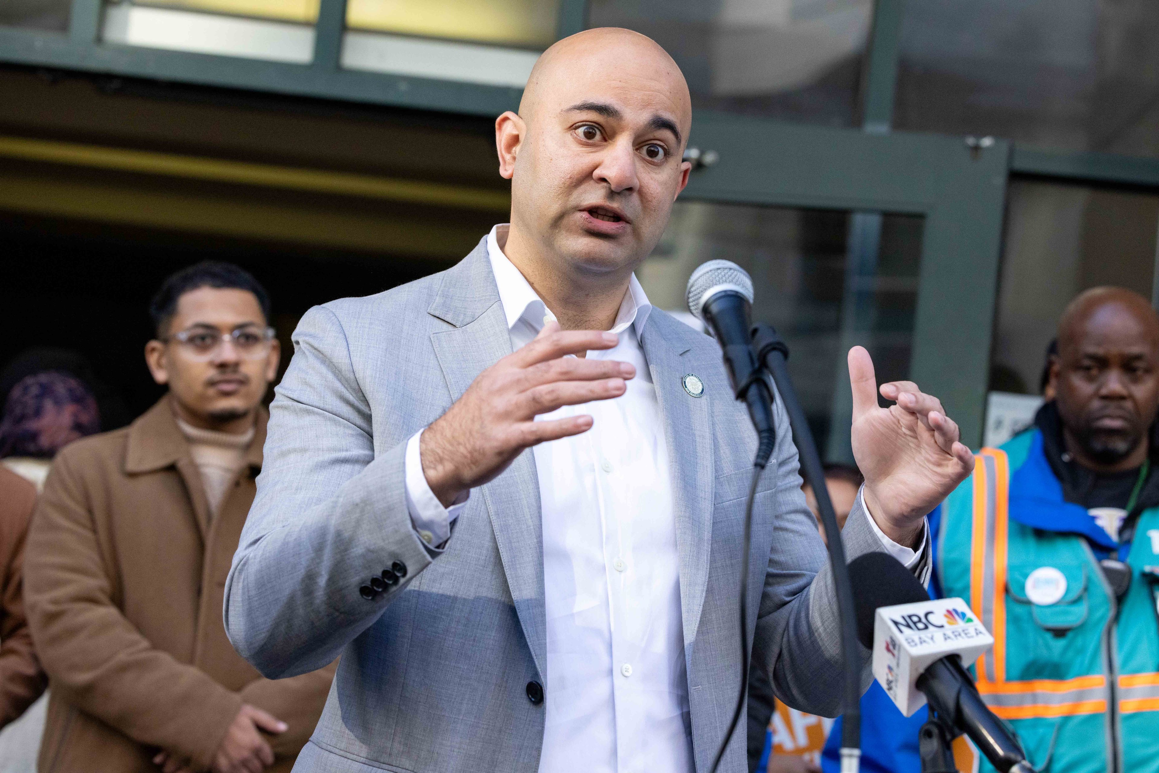 A man in a gray suit speaks passionately at a microphone with an &quot;NBC Bay Area&quot; sign. Two other men in the background observe him attentively.