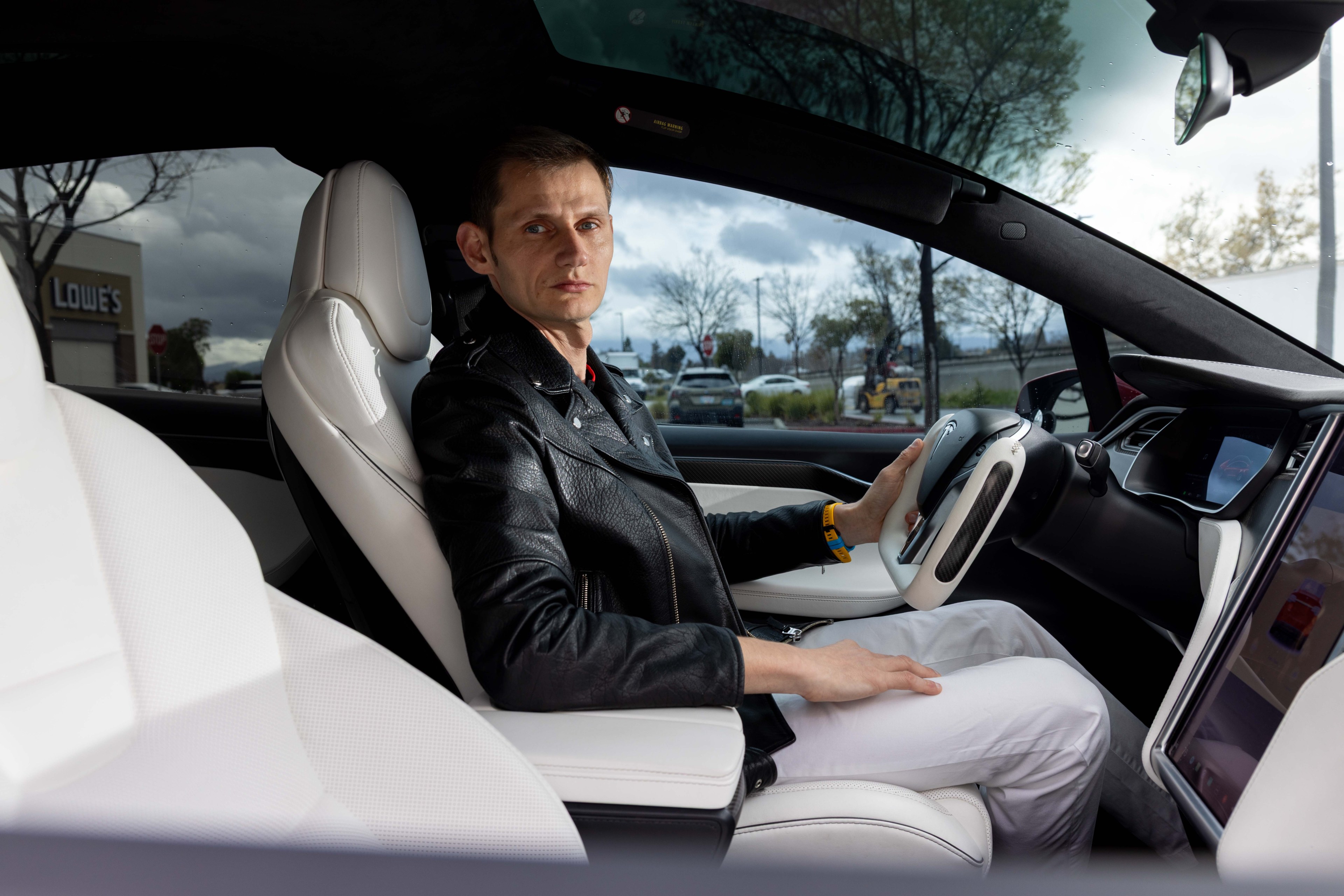 A person in a black leather jacket sits in the driver's seat of a Tesla with a confident expression. The interior features white seats and a large screen.