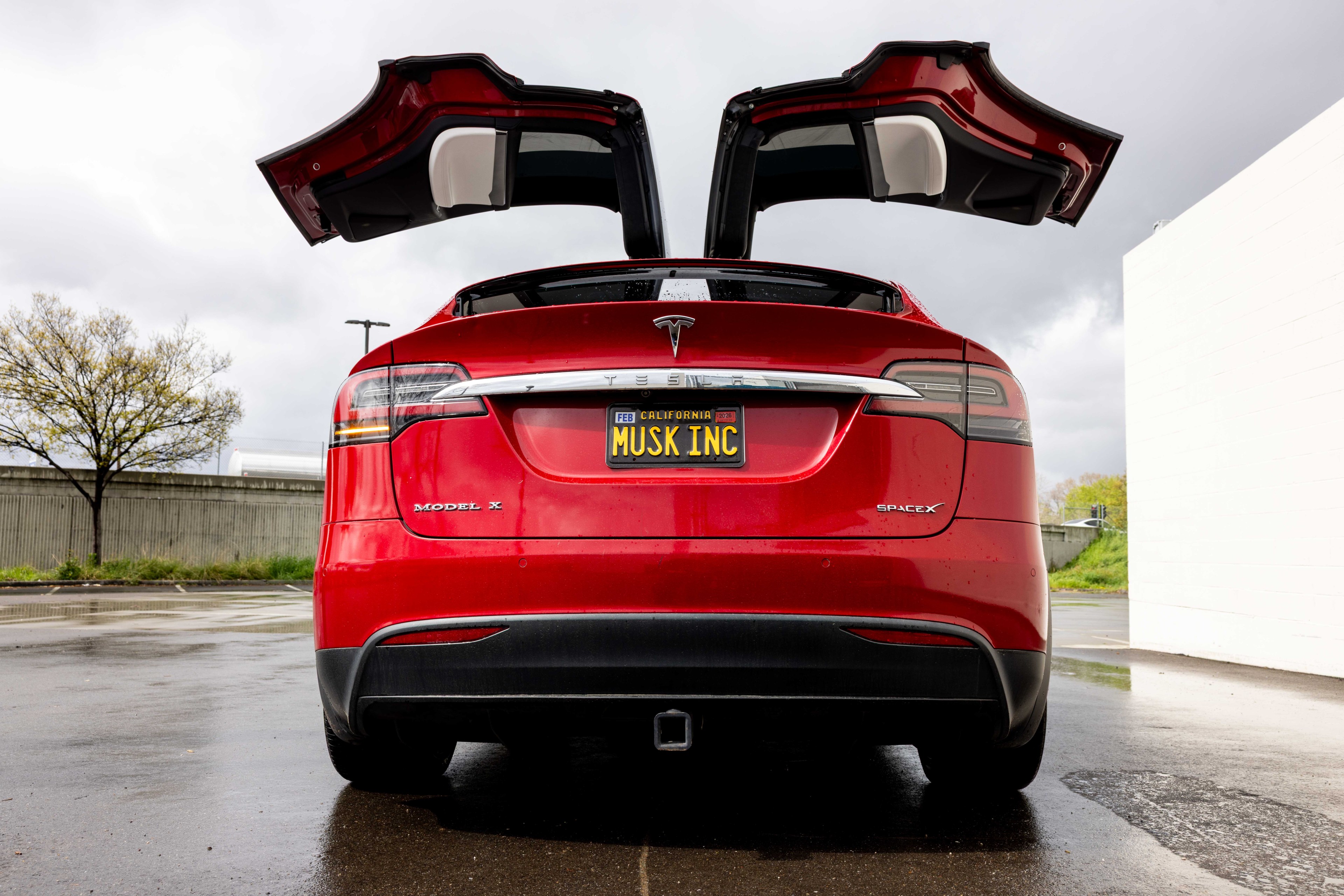 A red Tesla Model X with its rear gull-wing doors open is parked on a wet surface. The license plate reads &quot;MUSK INC,&quot; with trees and overcast sky in the background.