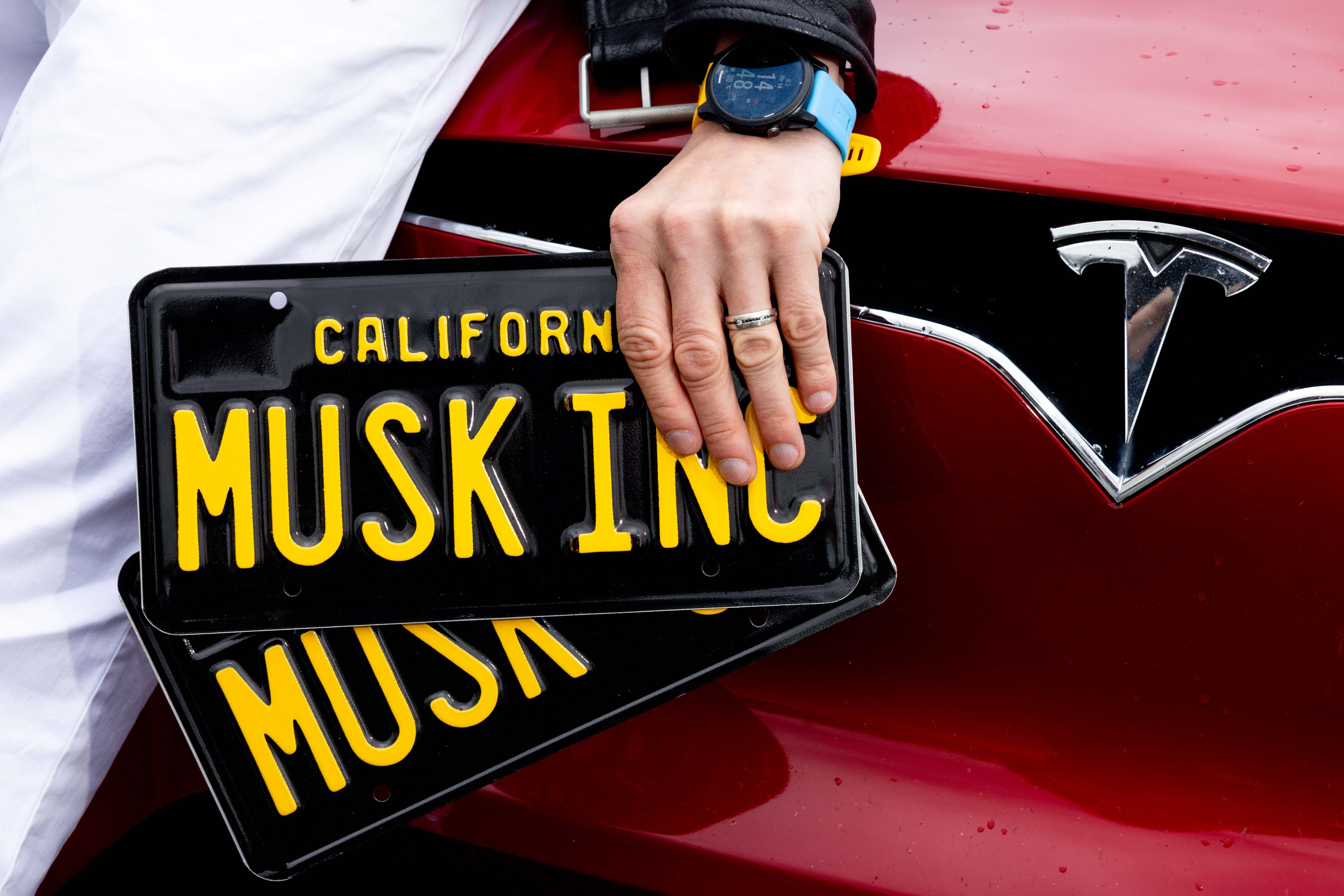 A hand rests on a red Tesla with two black California plates reading &quot;MUSK INC.&quot; The person has a smartwatch and is dressed in a black jacket.