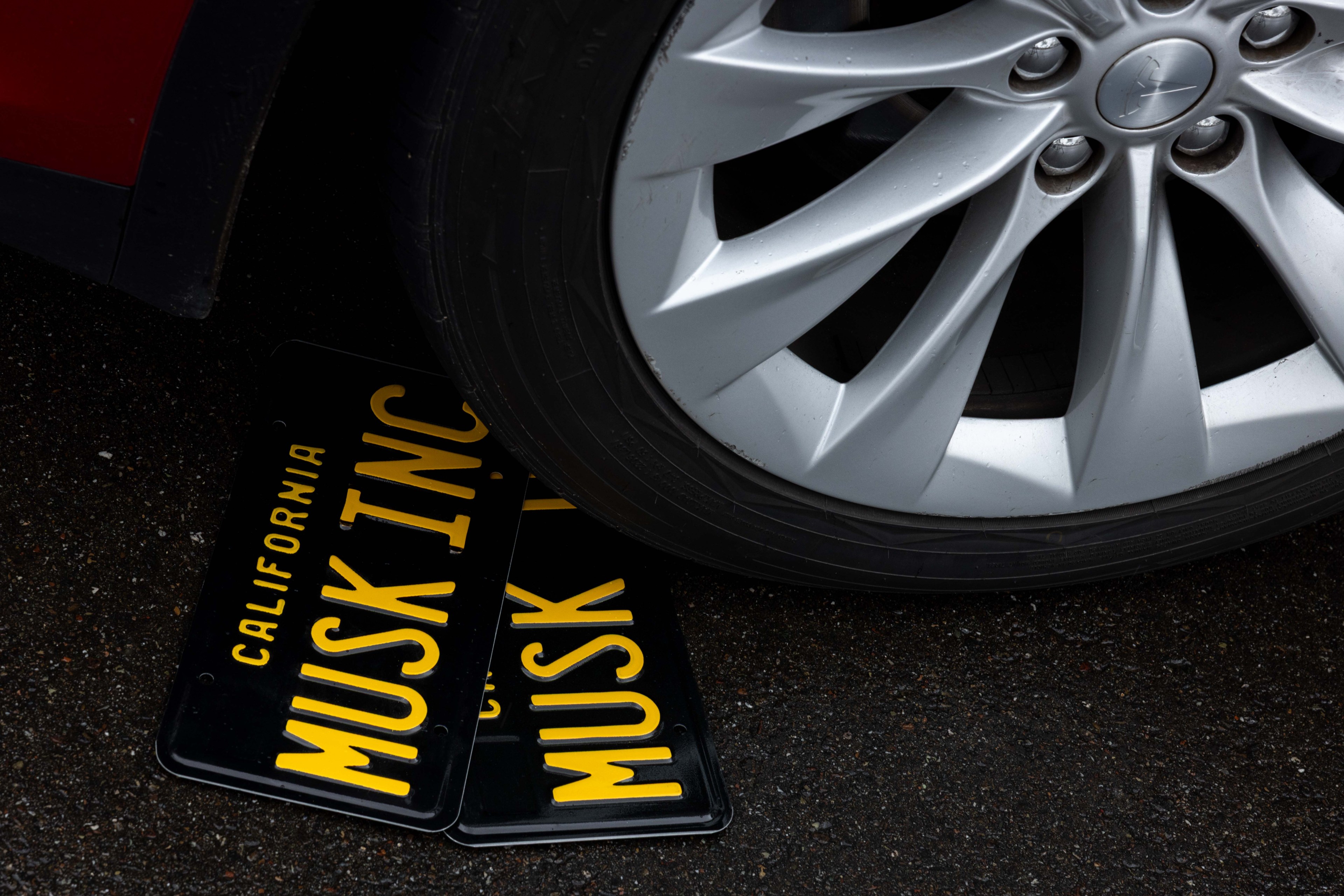 A silver car wheel partially rests on two black California license plates with yellow lettering saying &quot;MUSK INC.&quot; The ground is dark and wet.