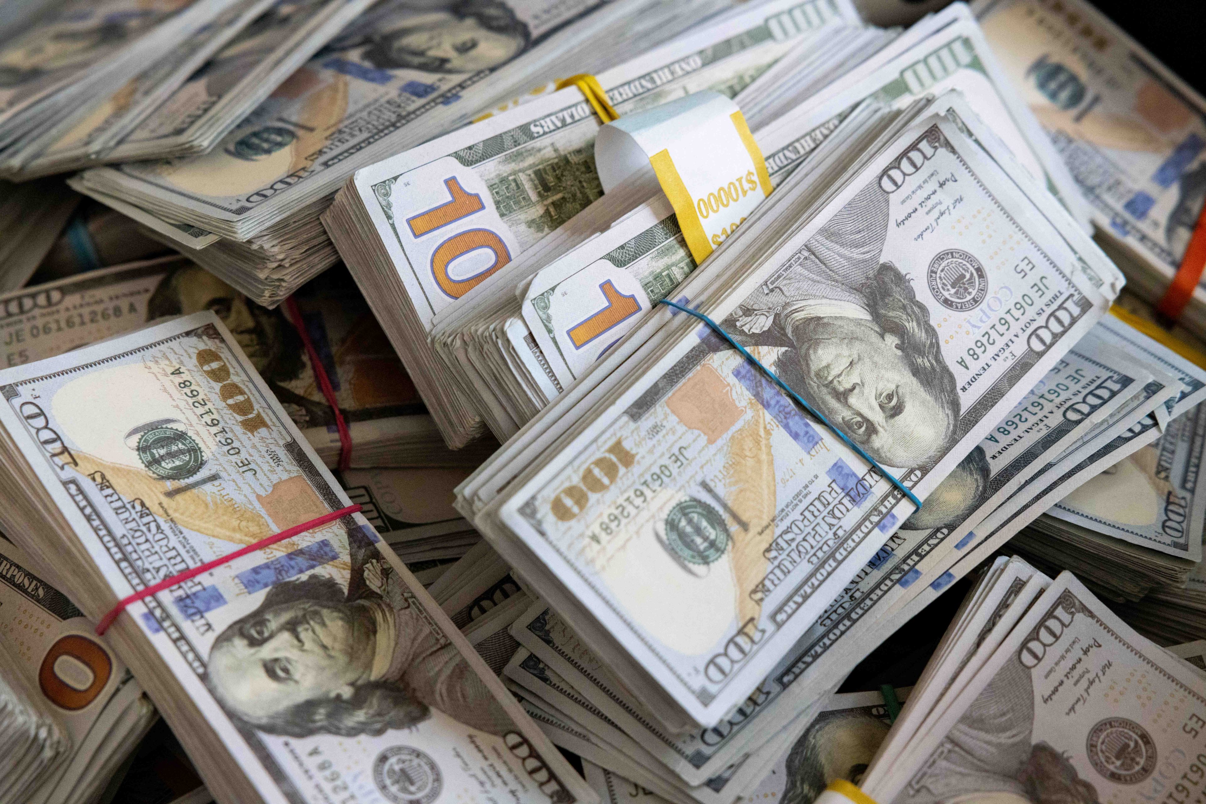 Bundles of false U.S. hundred-dollar bills are stacked closely together, secured with colored bands, showcasing piles of currency in a cluttered arrangement.