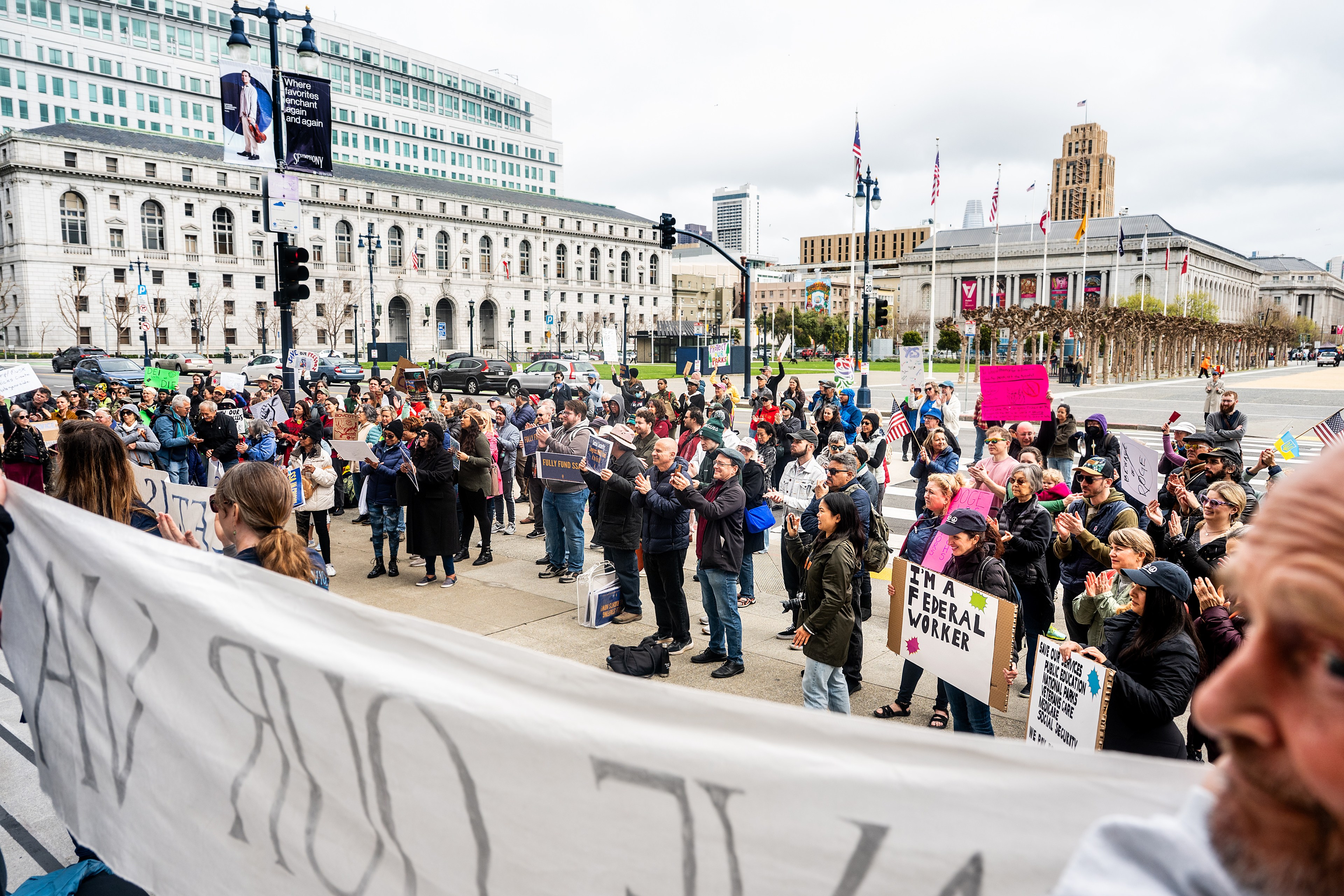 A large crowd gathers for a protest outside a government building, holding signs and applauding. The scene is dynamic with diverse participants.