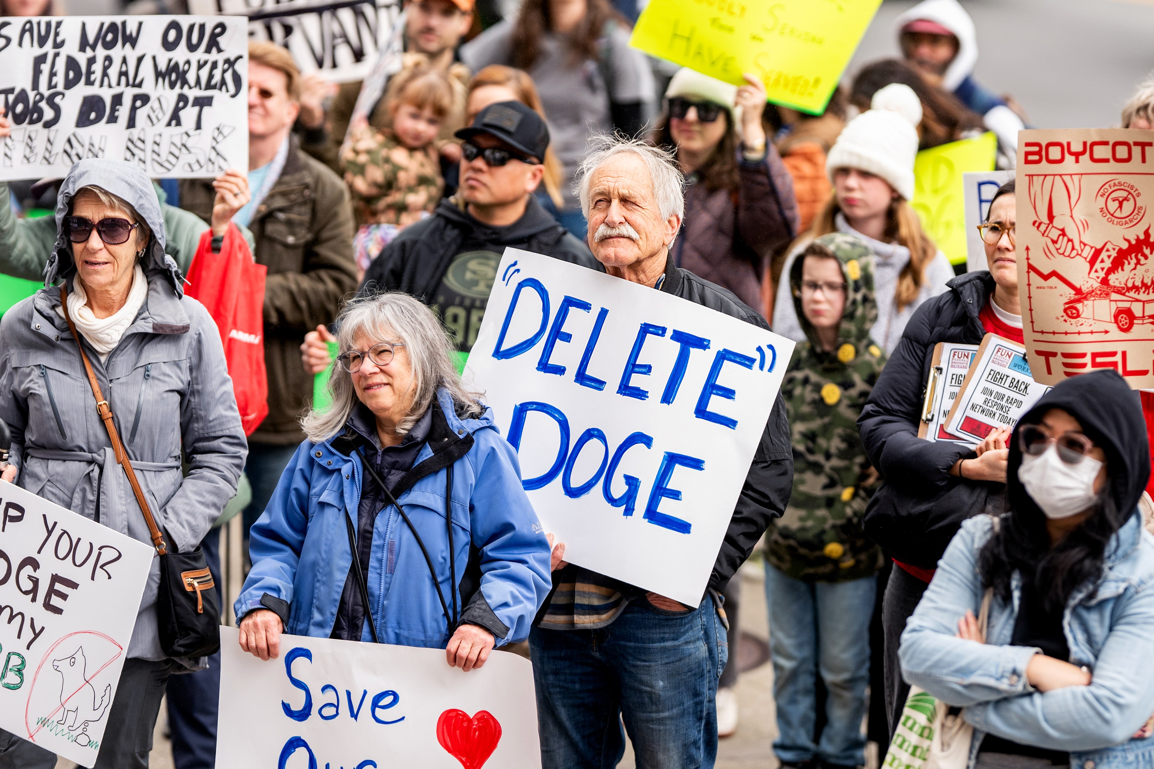 The image shows a crowd of protesters holding signs, including "Delete Doge" and "Boycott Tesla," expressing diverse social and political concerns.