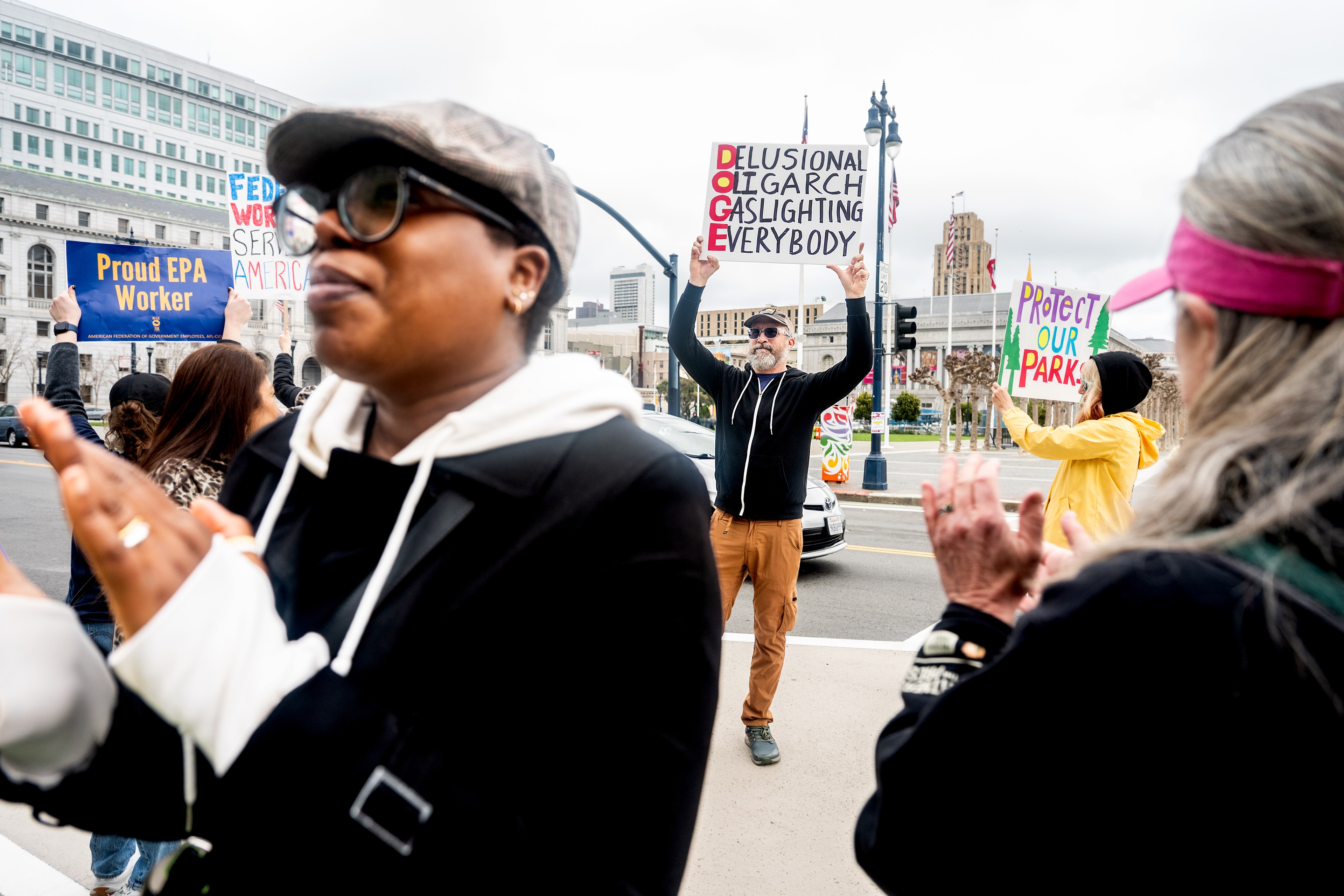 People are protesting outside, holding signs with messages like &quot;Proud EPA Worker&quot; and &quot;Protect Our Park.&quot; One person holds a sign criticizing a &quot;delusional oligarch.&quot;