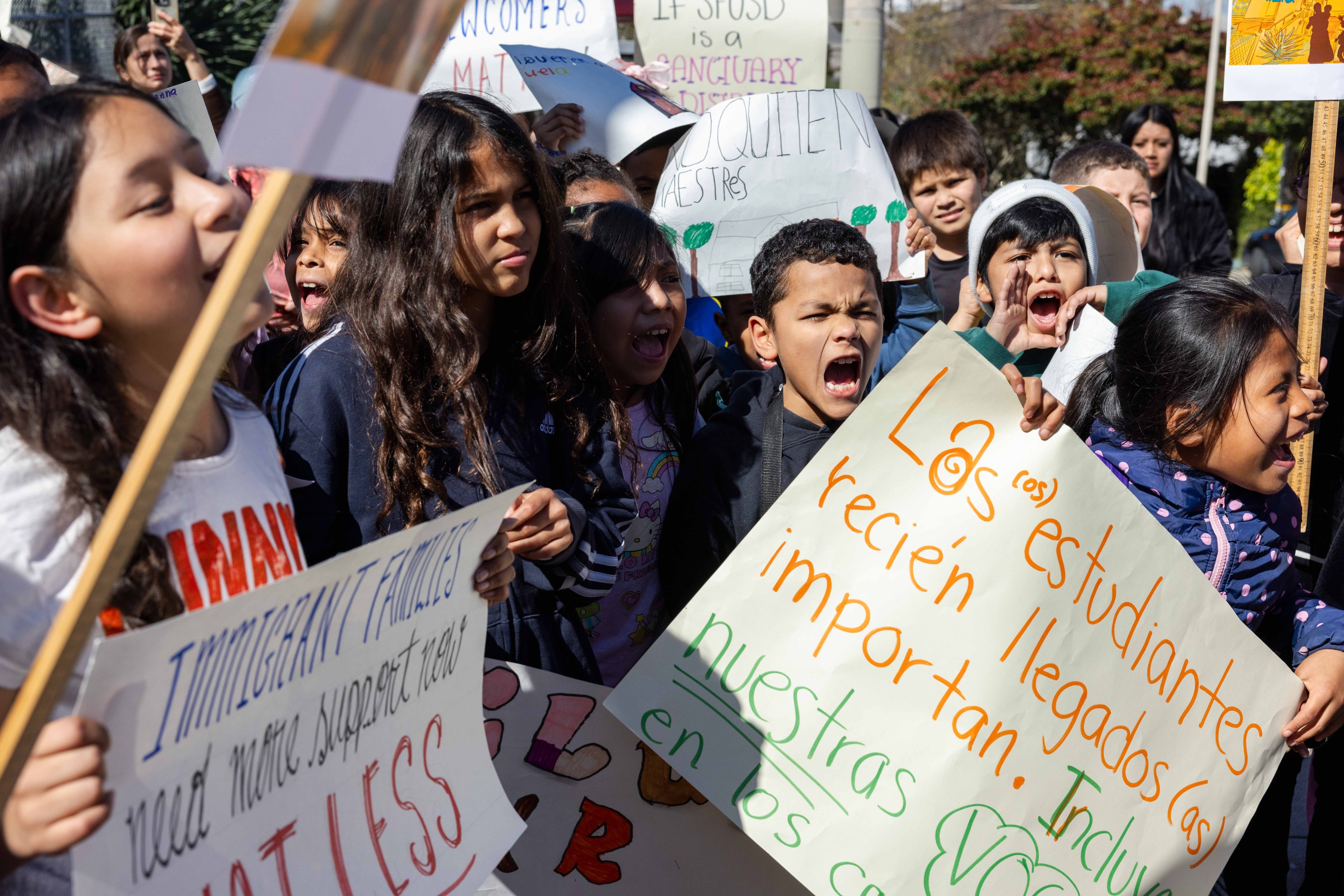 A group of children holds protest signs about supporting immigrant families and students. They appear passionate and vocal, with several signs in English and Spanish.