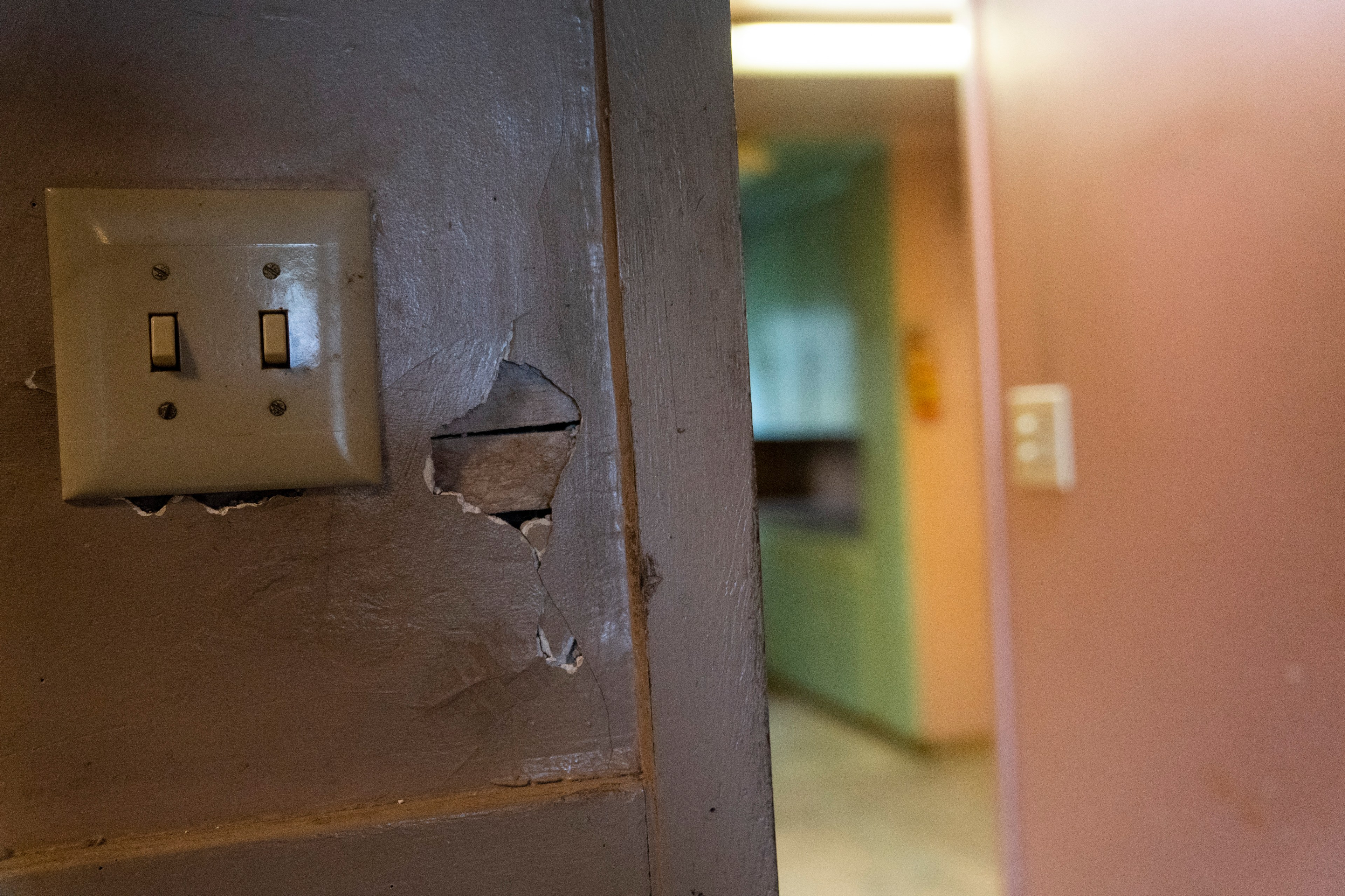 A damaged light switch plate on a cracked wall is in focus. In the background, there's a dimly lit, blurry view of a hallway or room.