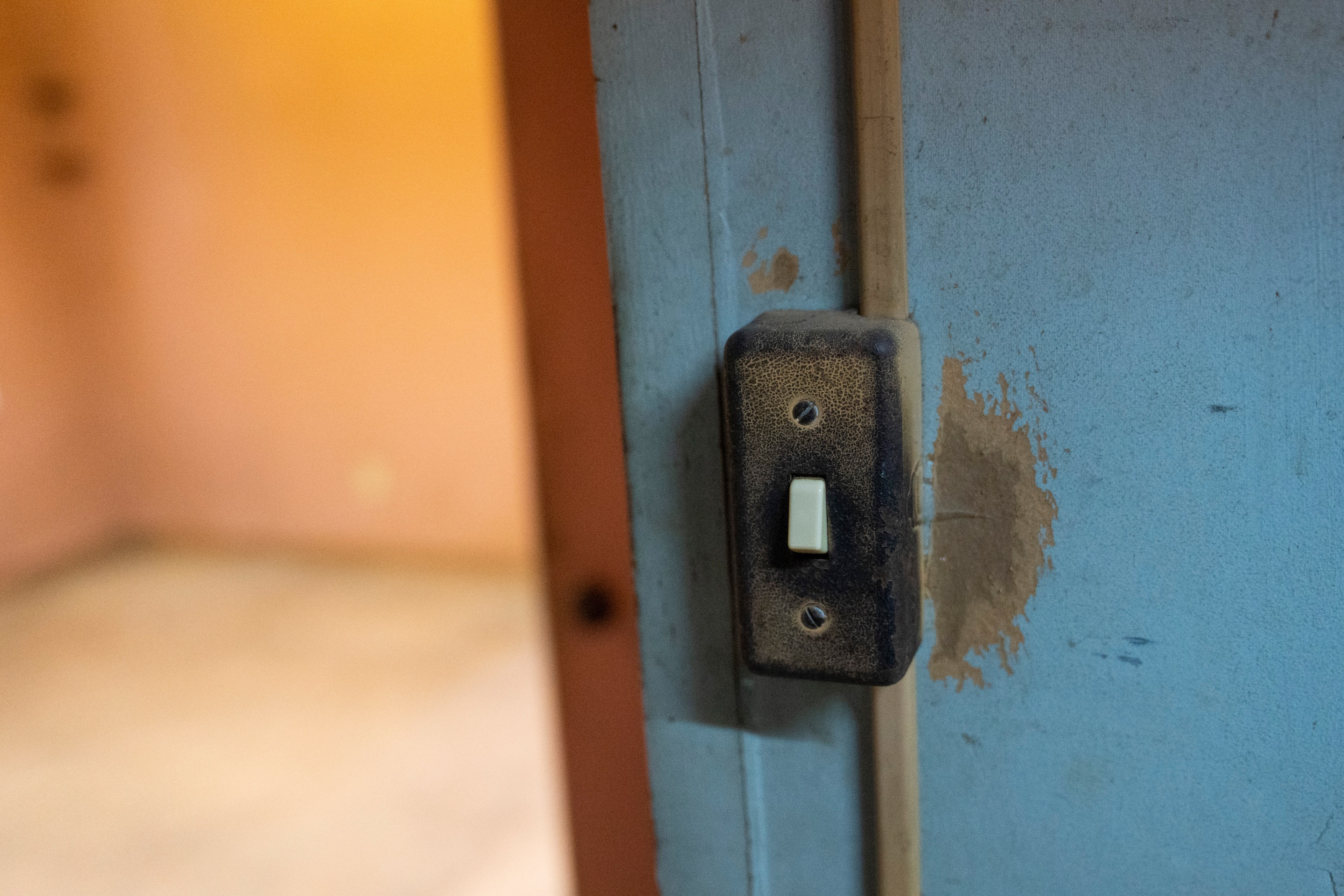 A dusty, worn light switch is attached to a blue wall with peeling paint, situated near a doorway leading to a dimly lit, beige-colored room.