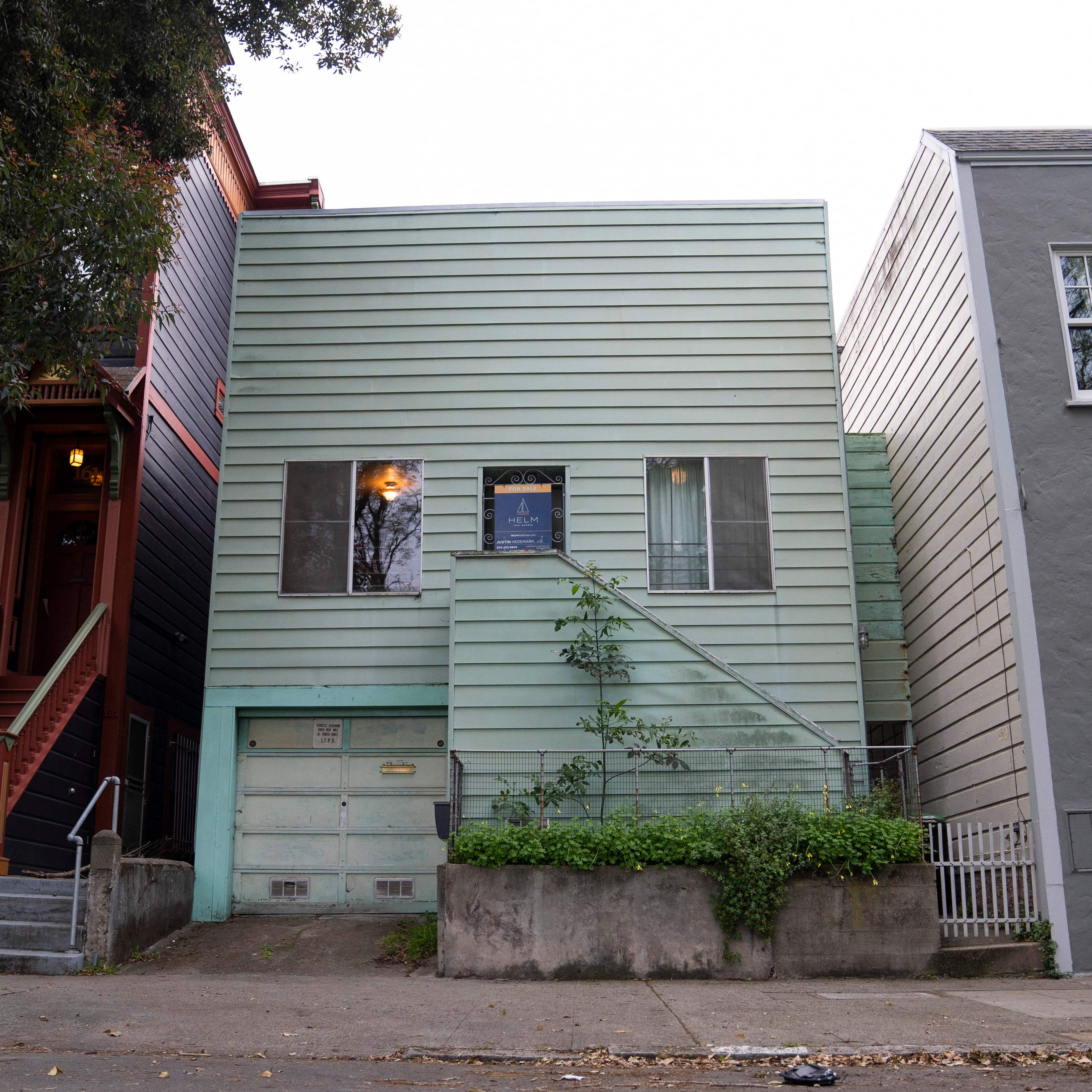 A small light green house with horizontal siding stands between two larger homes. It has a garage, two windows, and a simple staircase leading to the front door.