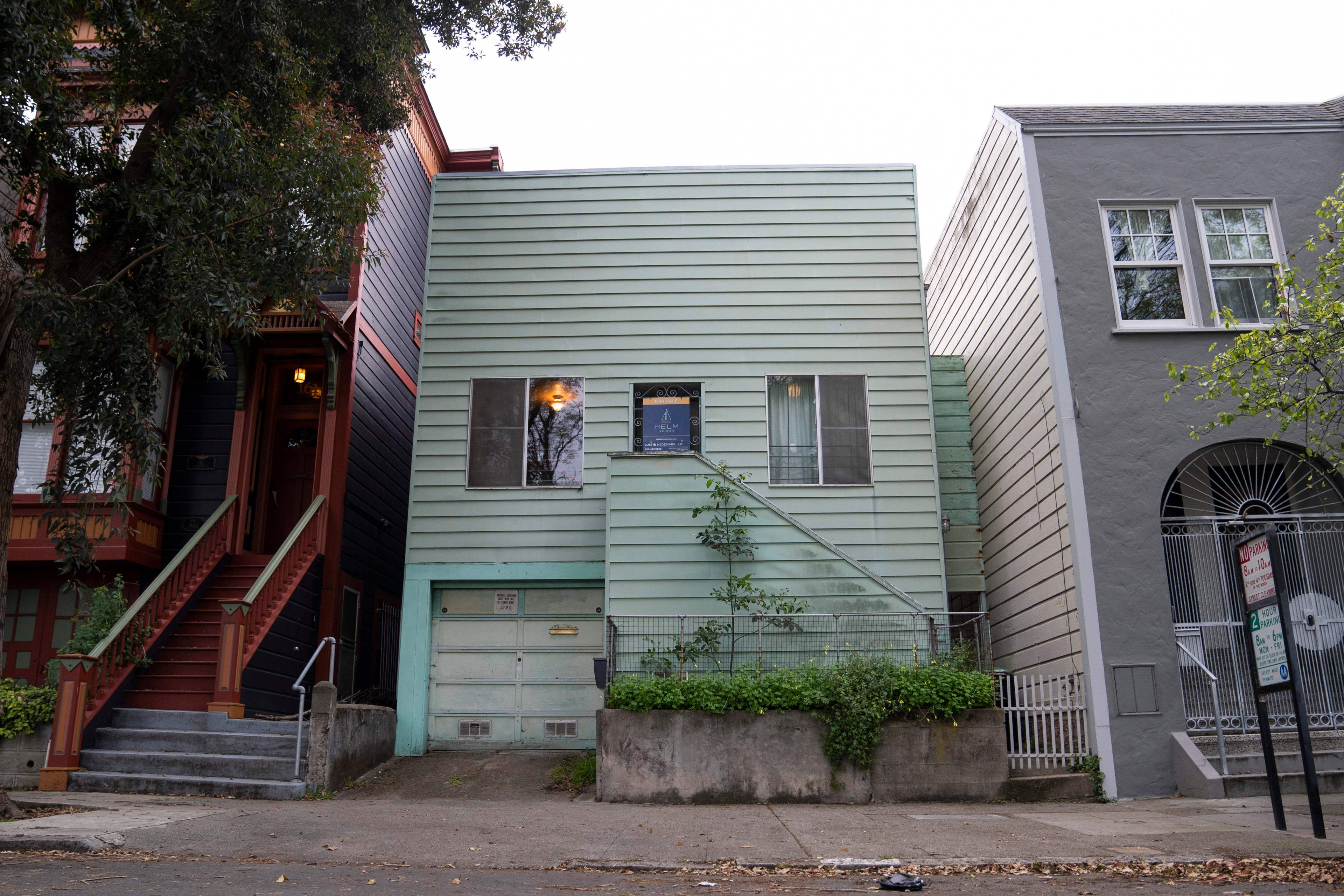 A small light green house with horizontal siding stands between two larger homes. It has a garage, two windows, and a simple staircase leading to the front door.