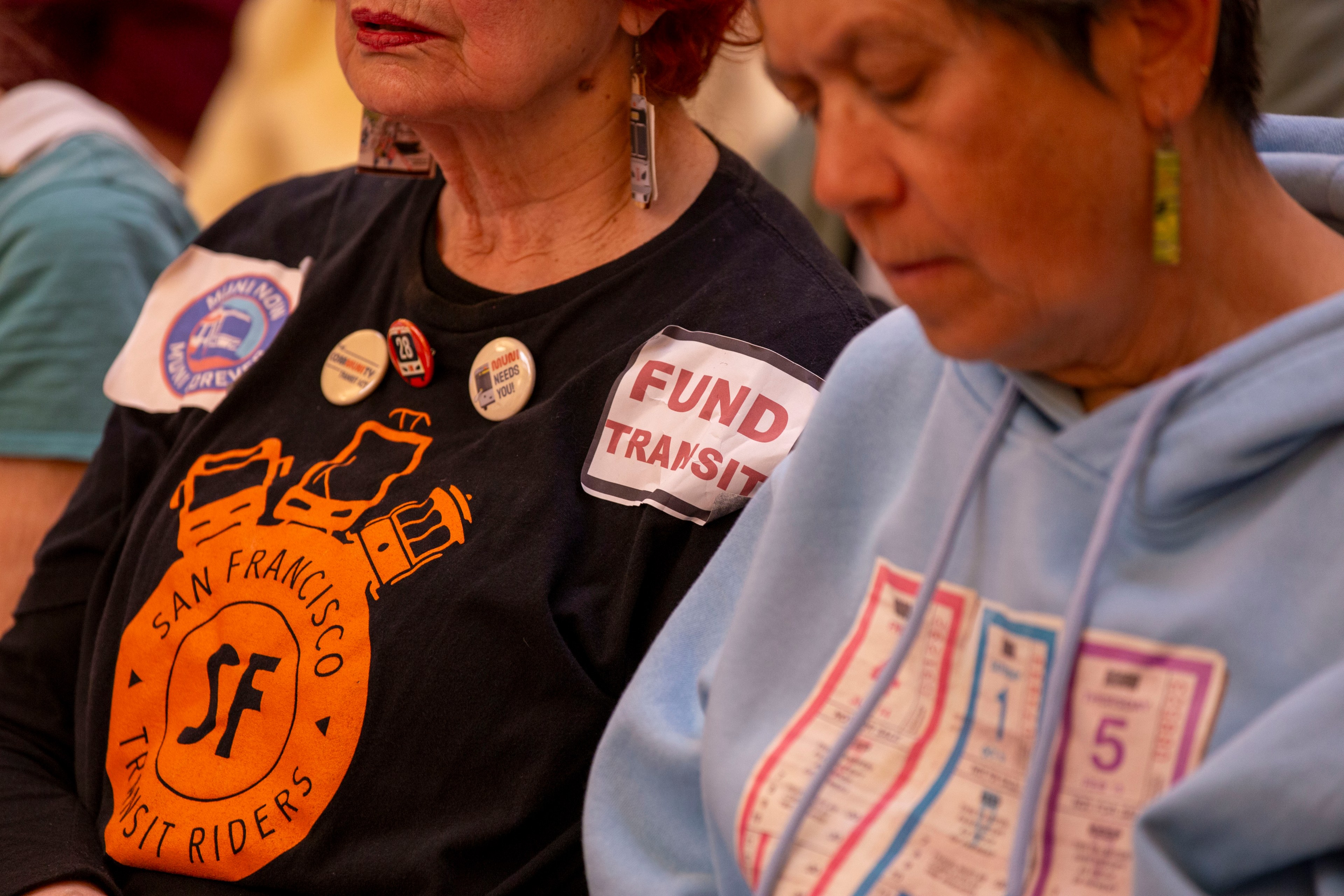 Two people are wearing transit-themed attire. One has a black shirt with &quot;San Francisco Transit Riders&quot; and buttons; the other wears a blue hoodie featuring ticket designs.