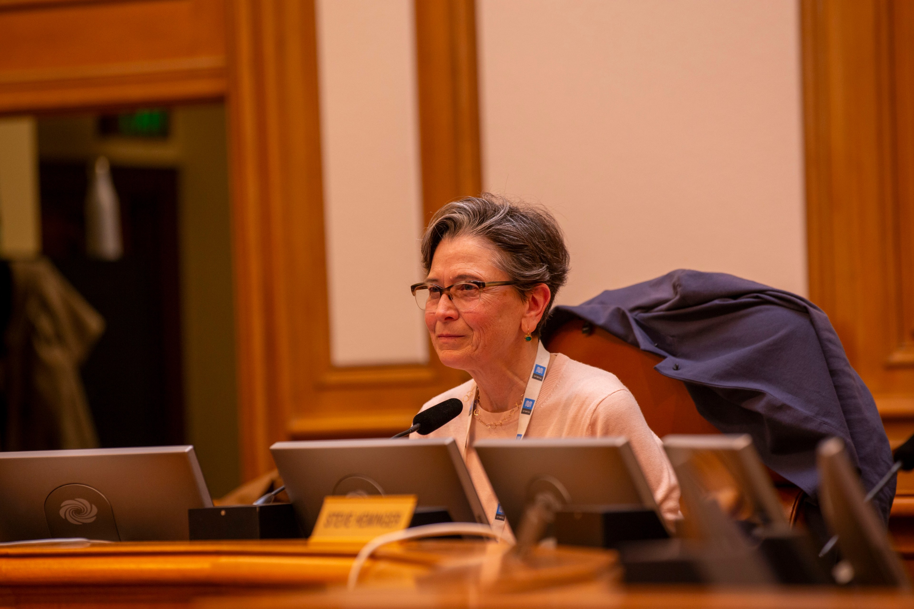 A person with short gray hair and glasses sits at a conference table with microphones and laptops, wearing a lanyard and a relaxed jacket over the chair.