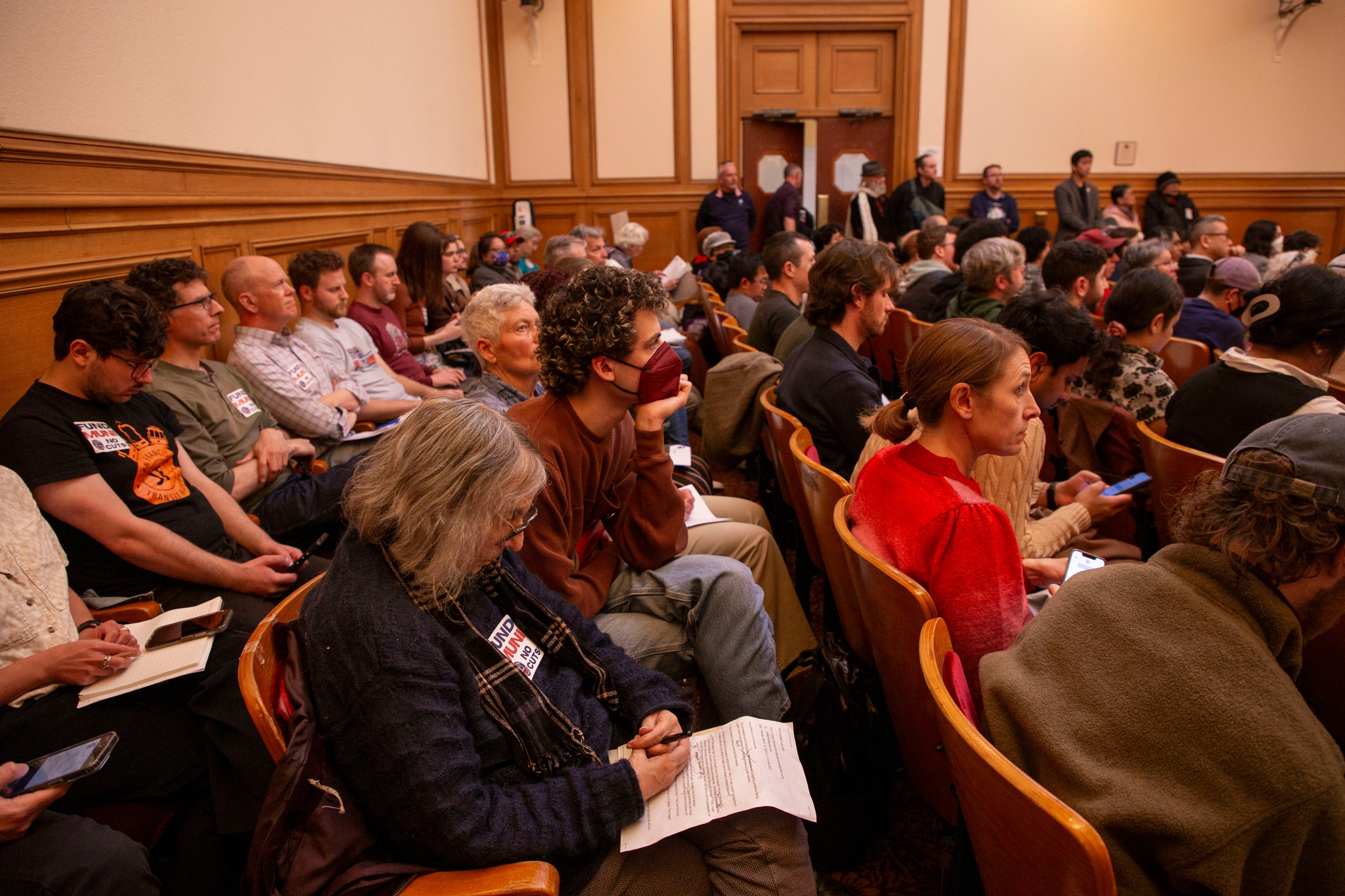 The image shows a packed meeting room with people seated closely together, some holding papers or using phones. A person in a mask is prominent in the middle.