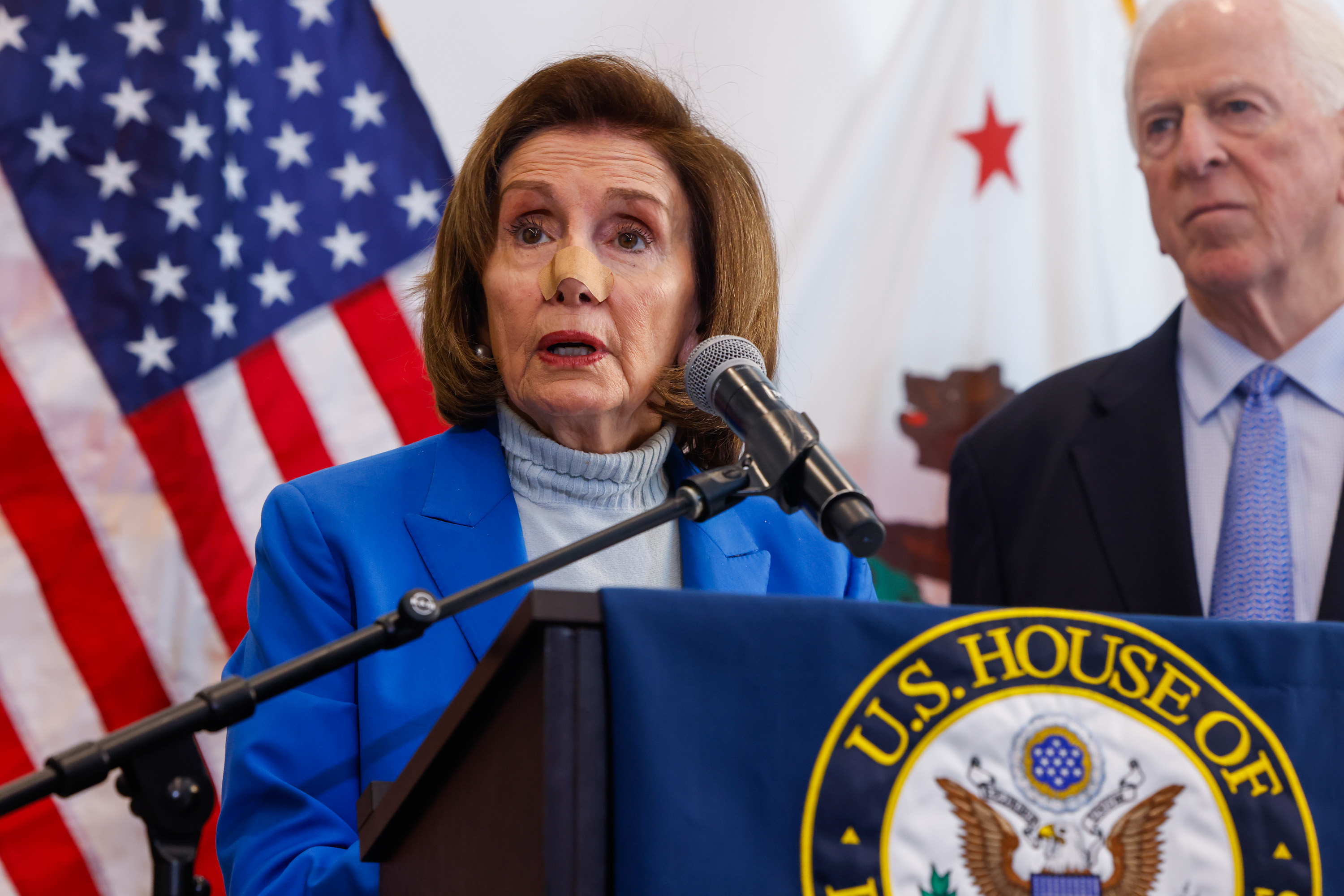 A woman in a blue outfit speaks at a podium with a microphone. An American flag and another person in a suit are in the background. A U.S. House banner is visible.