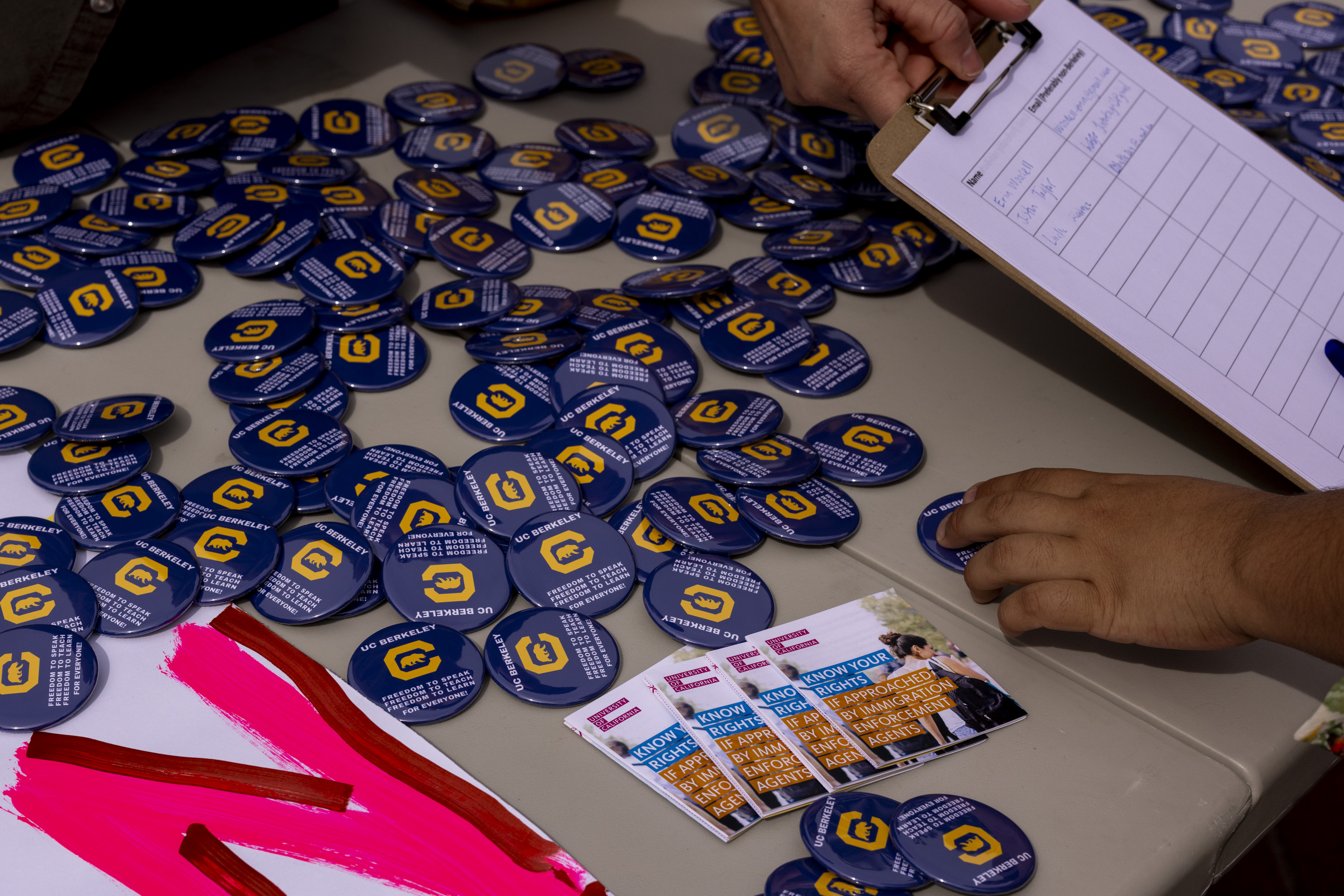 A table is covered with blue UC Berkeley buttons and informational pamphlets. A hand holds a clipboard with a signup sheet.