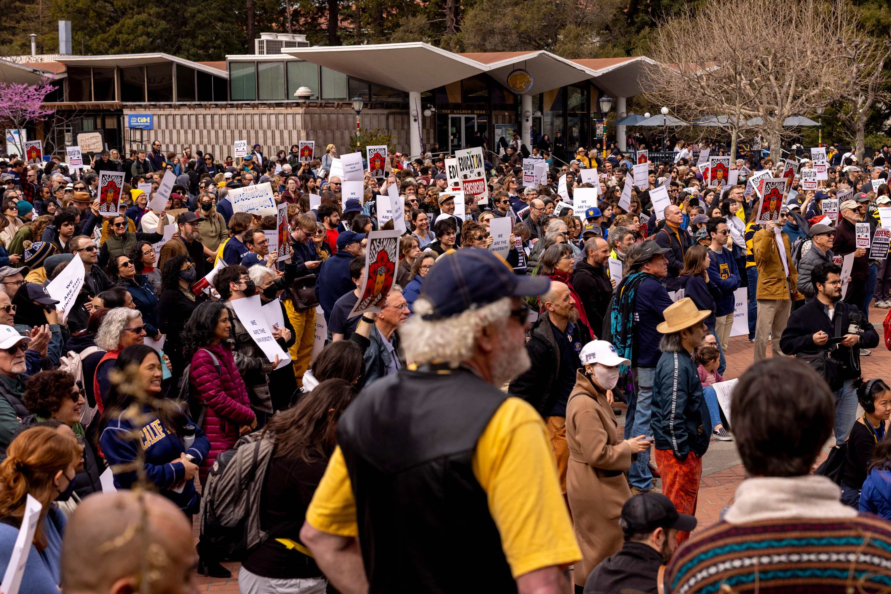 A large crowd gathers at a public square, holding various protest signs. The area is surrounded by trees and a modern building, signifying an organized demonstration.