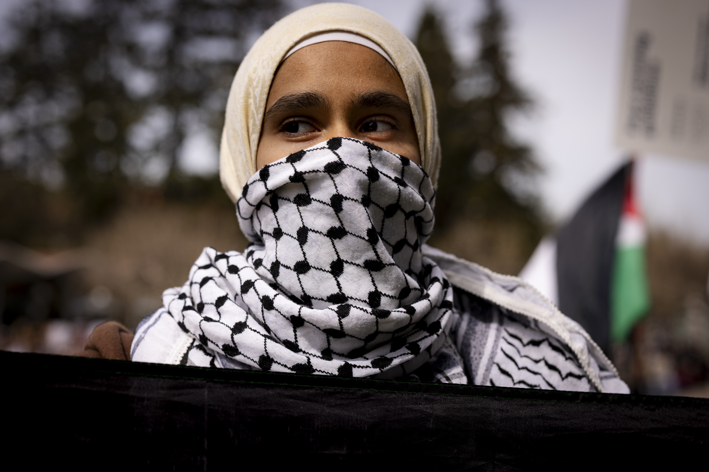 A person is wearing a beige headscarf and a black-and-white patterned scarf covering the lower face, with a blurry tree and flag background.