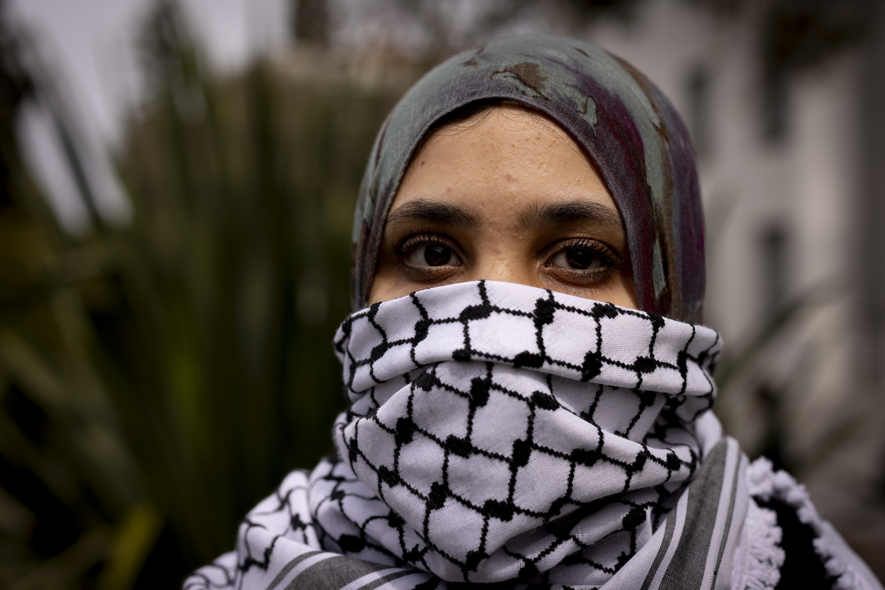 A person wearing a patterned hijab and a black and white checkered scarf covering their mouth and nose, with focus on their expressive eyes in a natural setting.