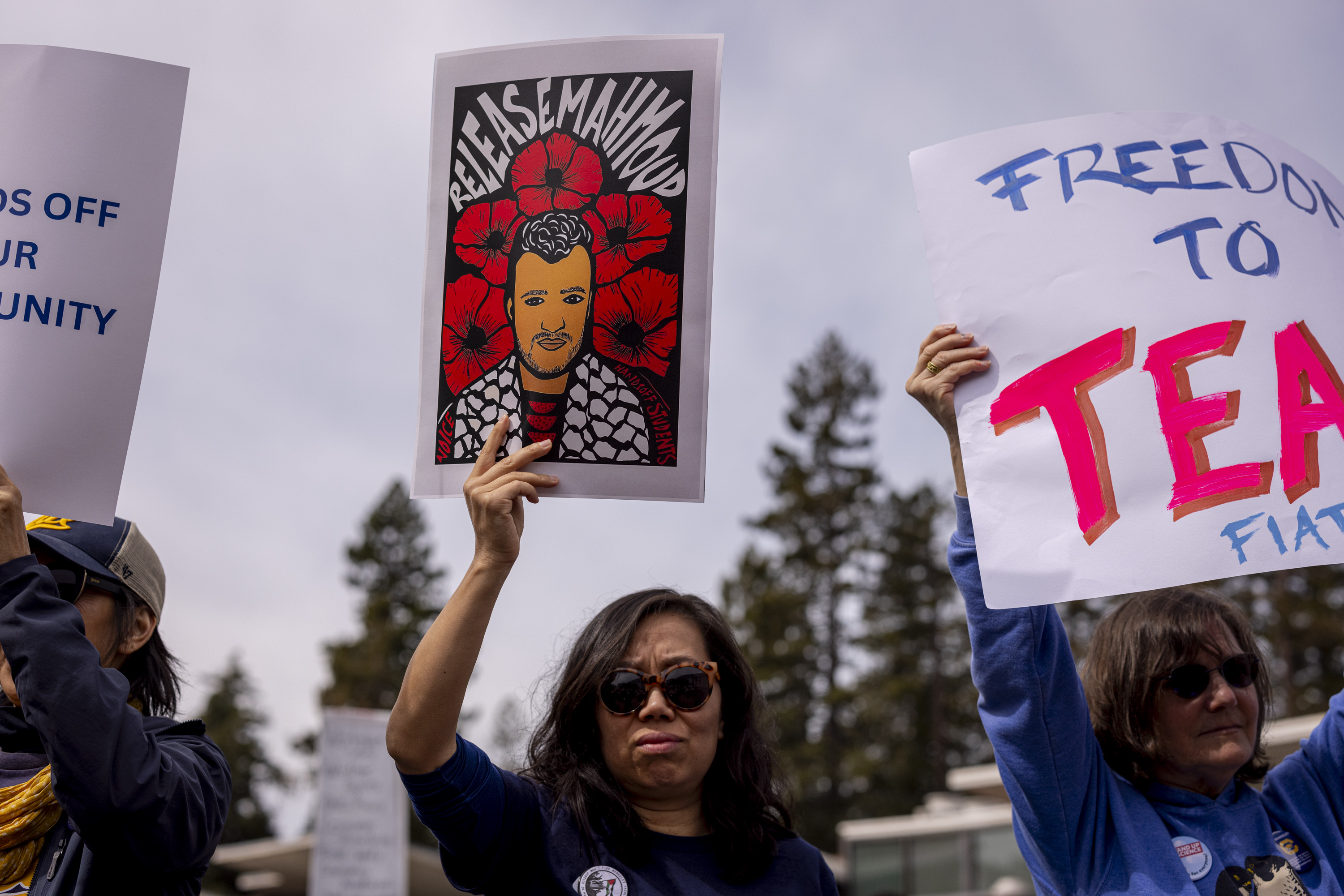 Protesters hold signs advocating for freedom, one depicting a person with flowers and text related to their release, amidst an outdoor setting with trees.