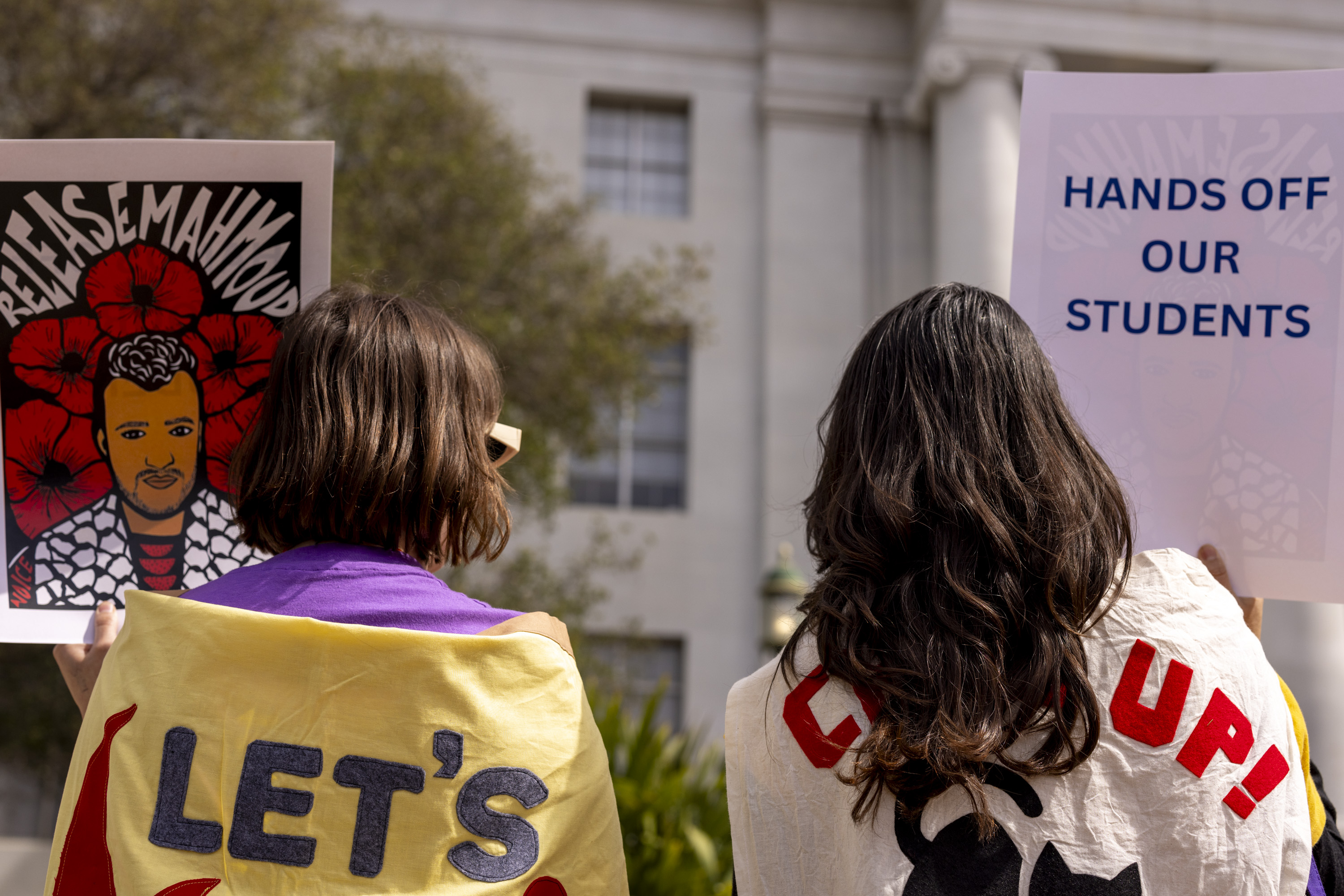 Two people hold protest signs: one features a portrait with &quot;RELEASE MAHMOUD,&quot; the other reads &quot;HANDS OFF OUR STUDENTS.&quot; They wear colorful cloaks.